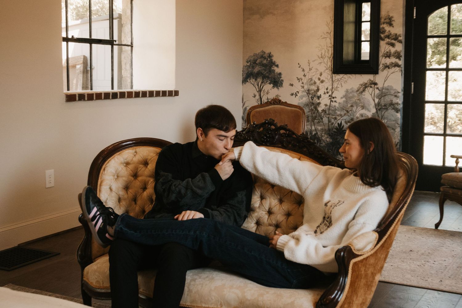 Couple sitting intimately on a vintage velvet sofa in an elegant room with scenic wallpaper and natural light.