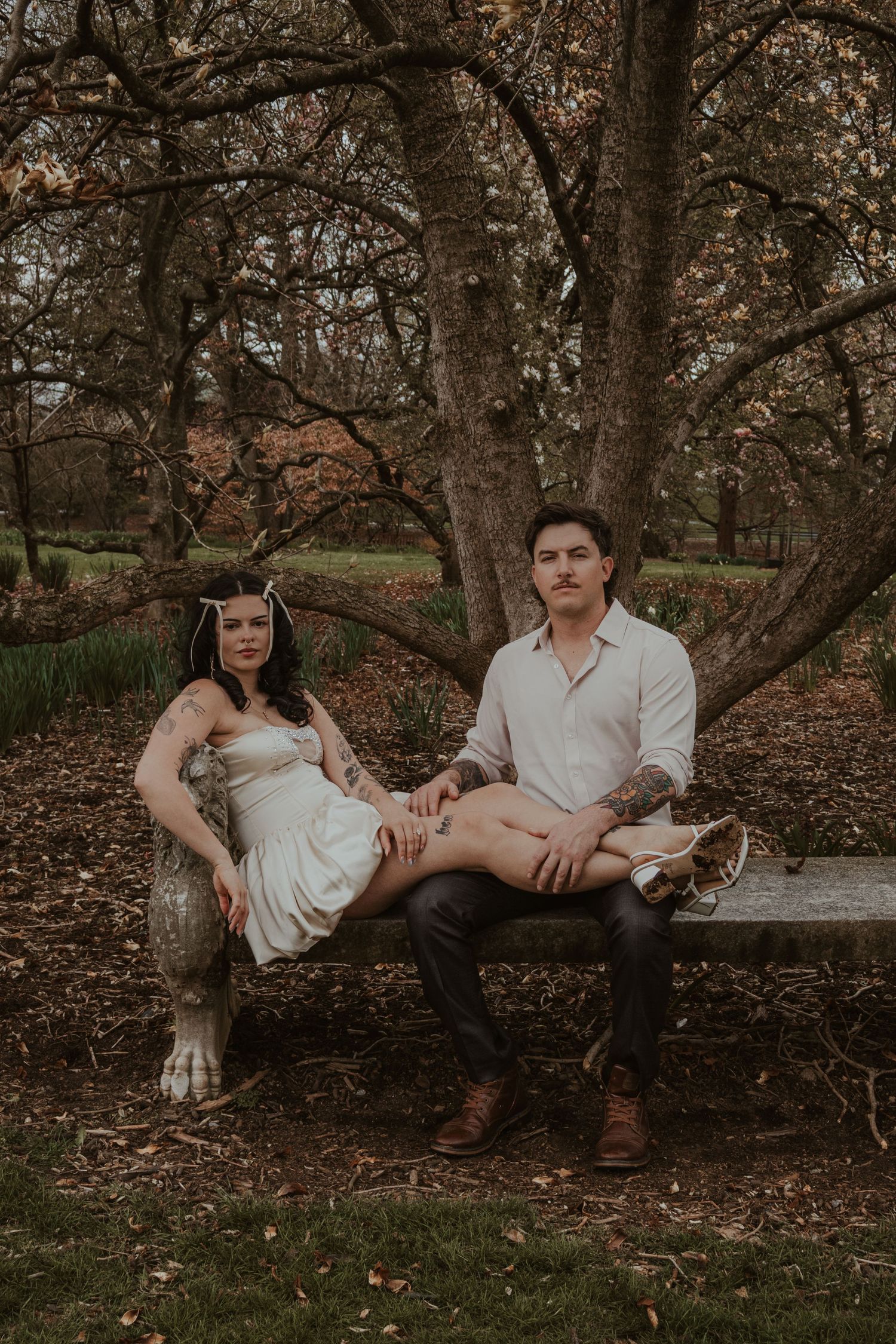 Couple with tattoos sitting on a stone bench under bare trees in a park, woman in white dress with floral headpiece.