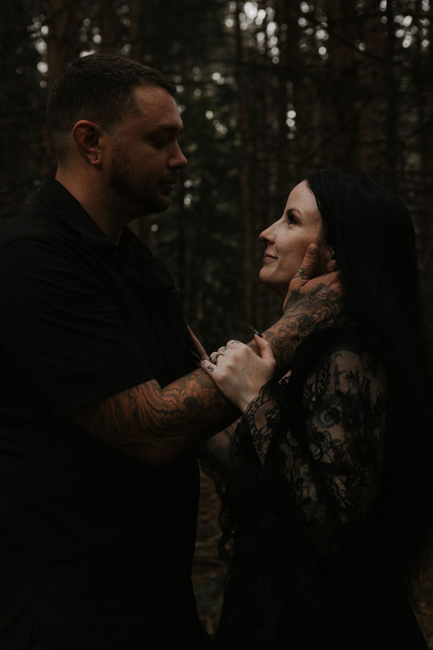 Tattooed couple in black outfits share a romantic moment in a dark forest, man tenderly holding womans face.