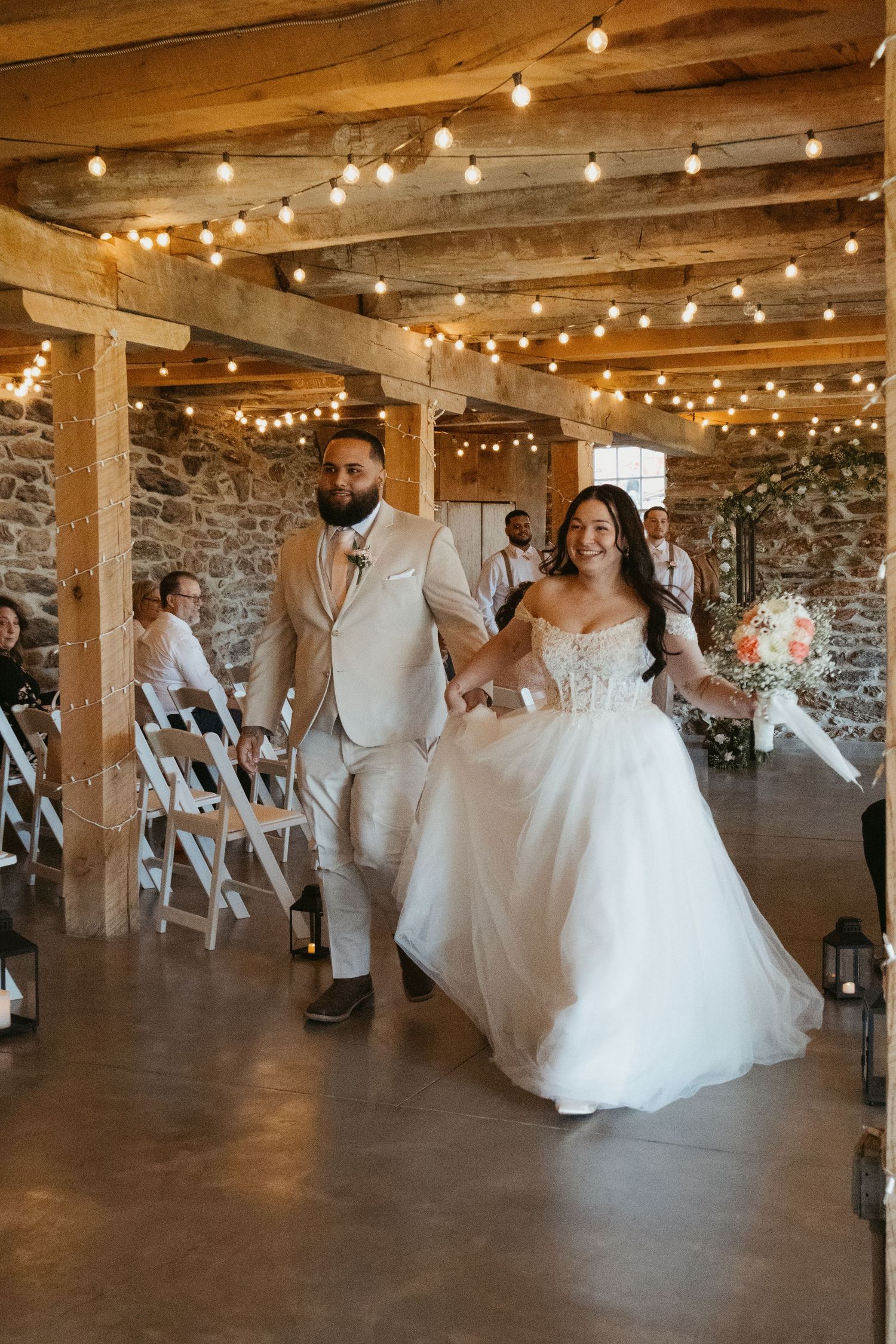 A joyful bride and groom walk hand-in-hand down the aisle in a rustic barn venue adorned with string lights.