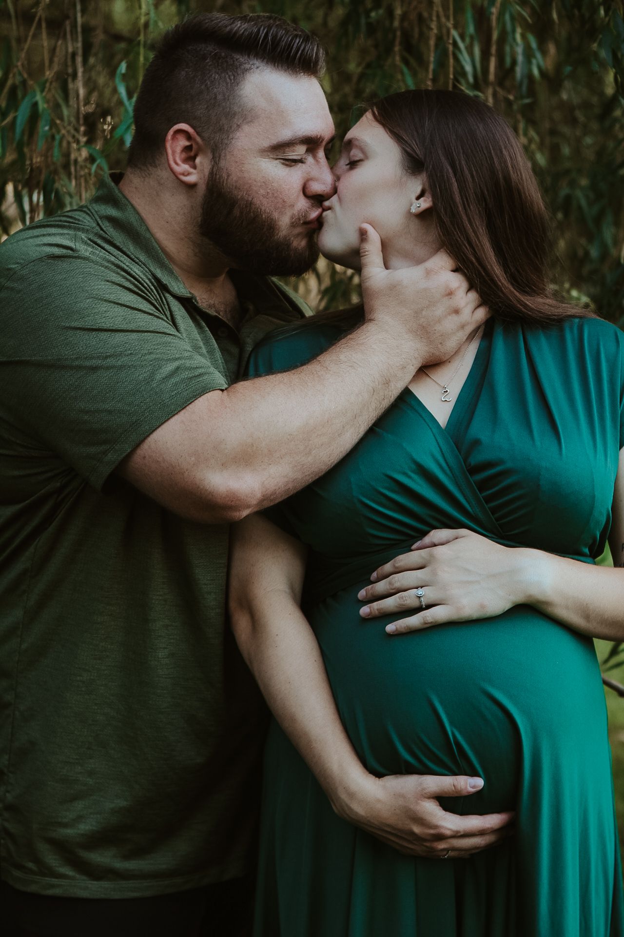 A couple in matching forest green clothing share an intimate moment during a maternity photoshoot in nature.