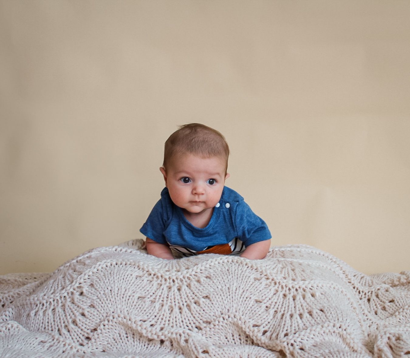 A baby in blue lies on a crocheted cream lace blanket while laying on their belly and looking at the camera.