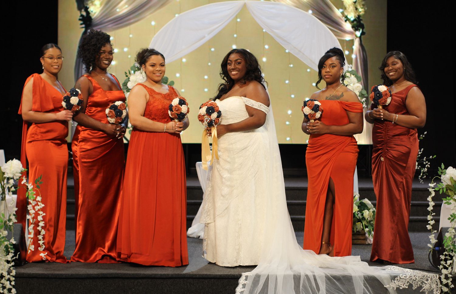 Wedding party in vibrant red dresses posing with bride in elegant white gown on stage.