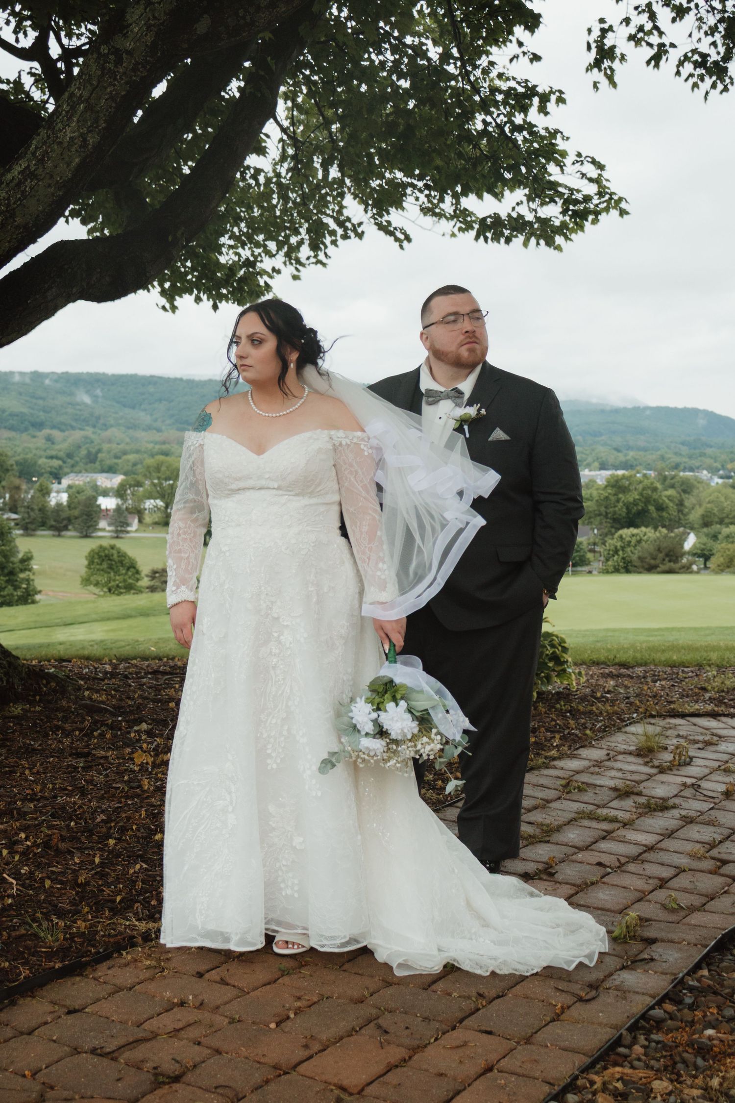 Bride in lace off-shoulder gown with veil and groom in black tux posing on brick path with scenic backdrop.