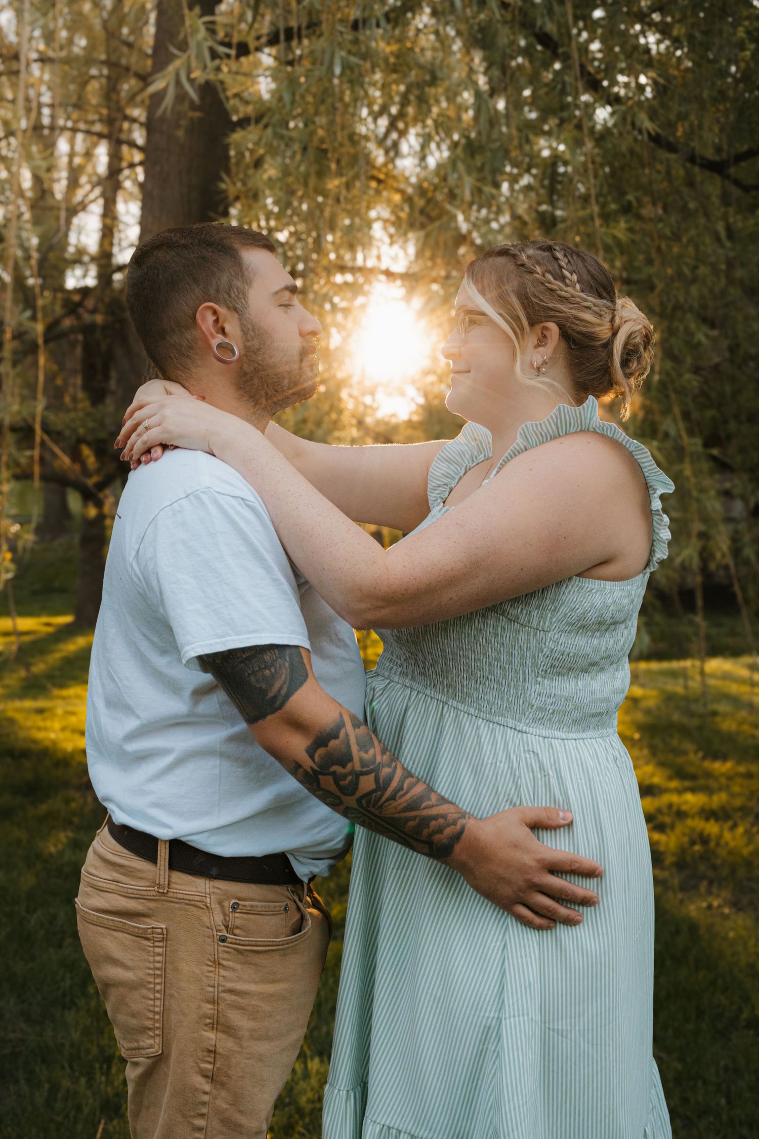 Couple embracing in golden sunset light in a lush green park, man with tattoos in white tee, woman in mint dress.