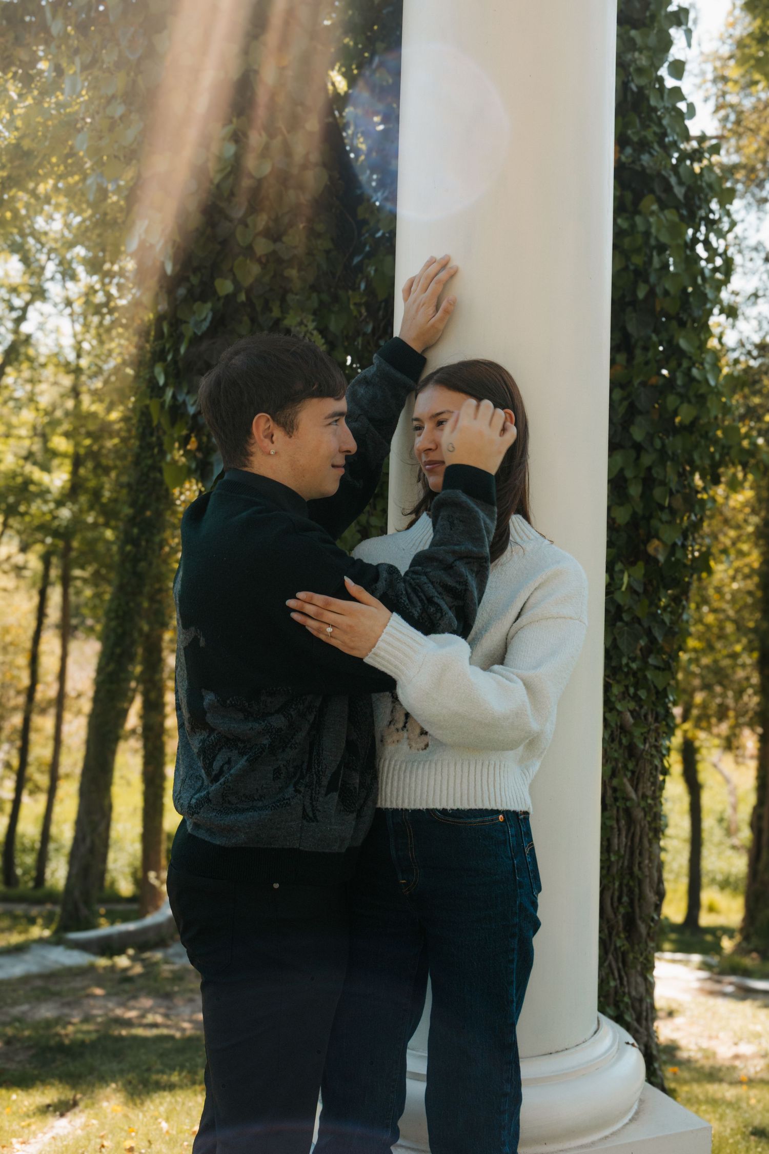 A young couple shares an intimate moment by a white column in a sunlit park, the woman leaning against it.