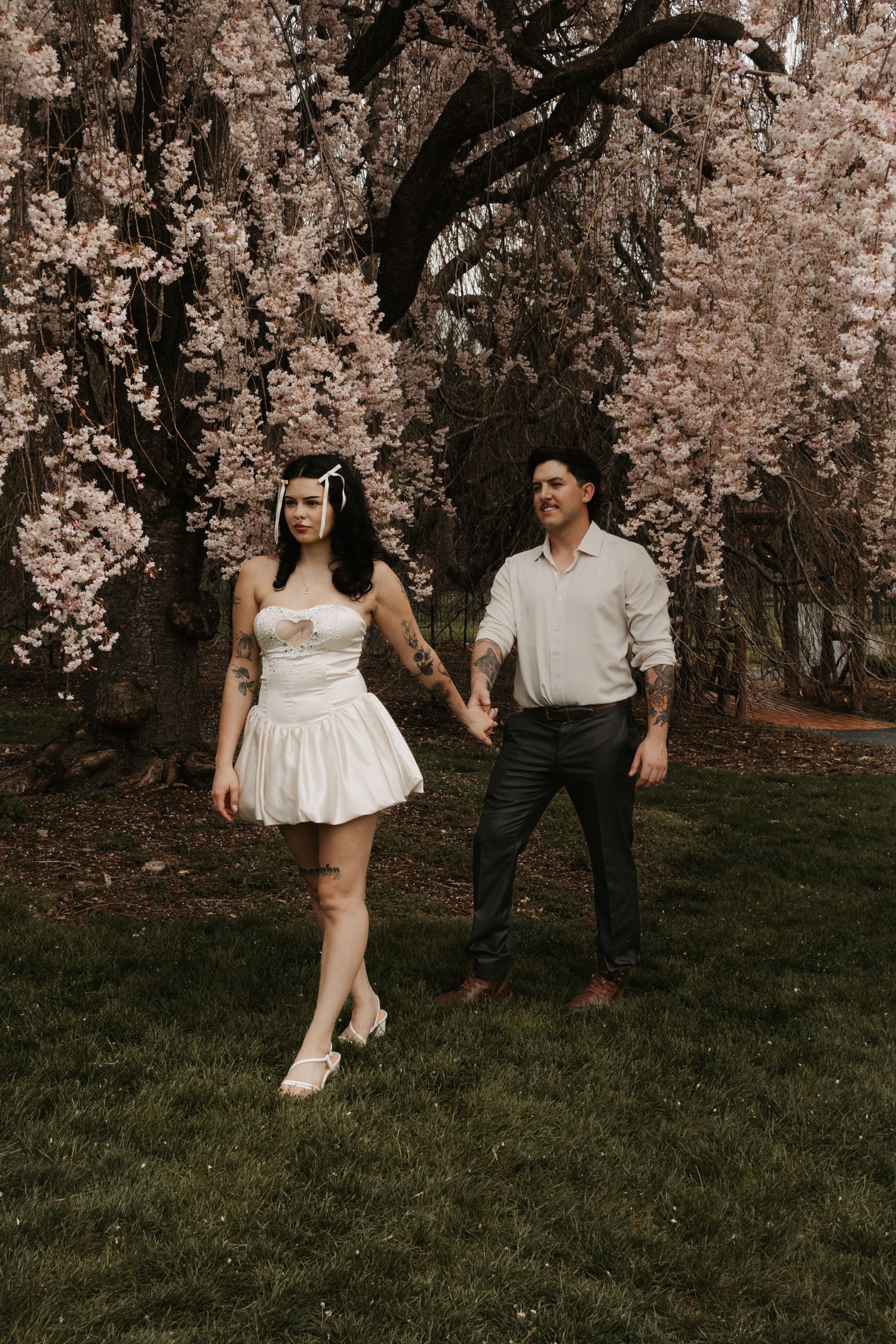 Couple holding hands under blooming cherry blossom tree, woman in white mini dress, man in light shirt and dark pants.