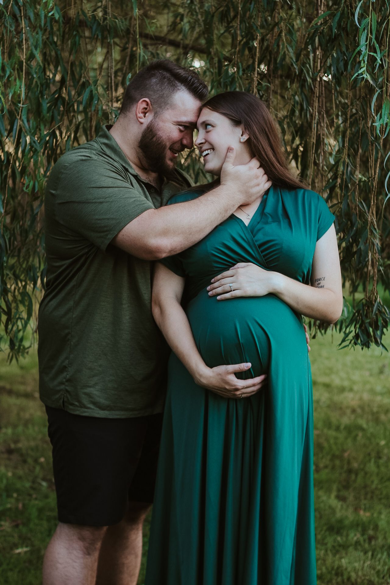 Romantic maternity portrait with couple laughing together against willow tree backdrop.