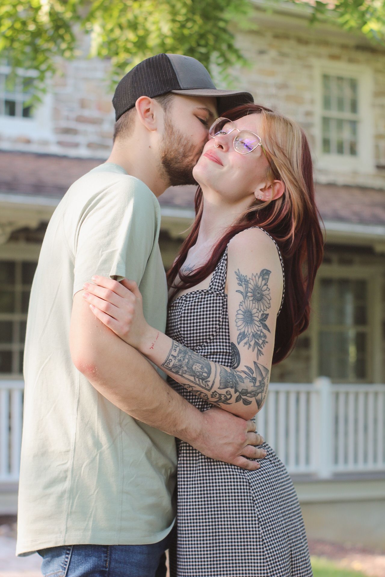 A couple embraces outdoors in front of a building, with one wearing a black cap and the other in a gingham dress with tattoos.