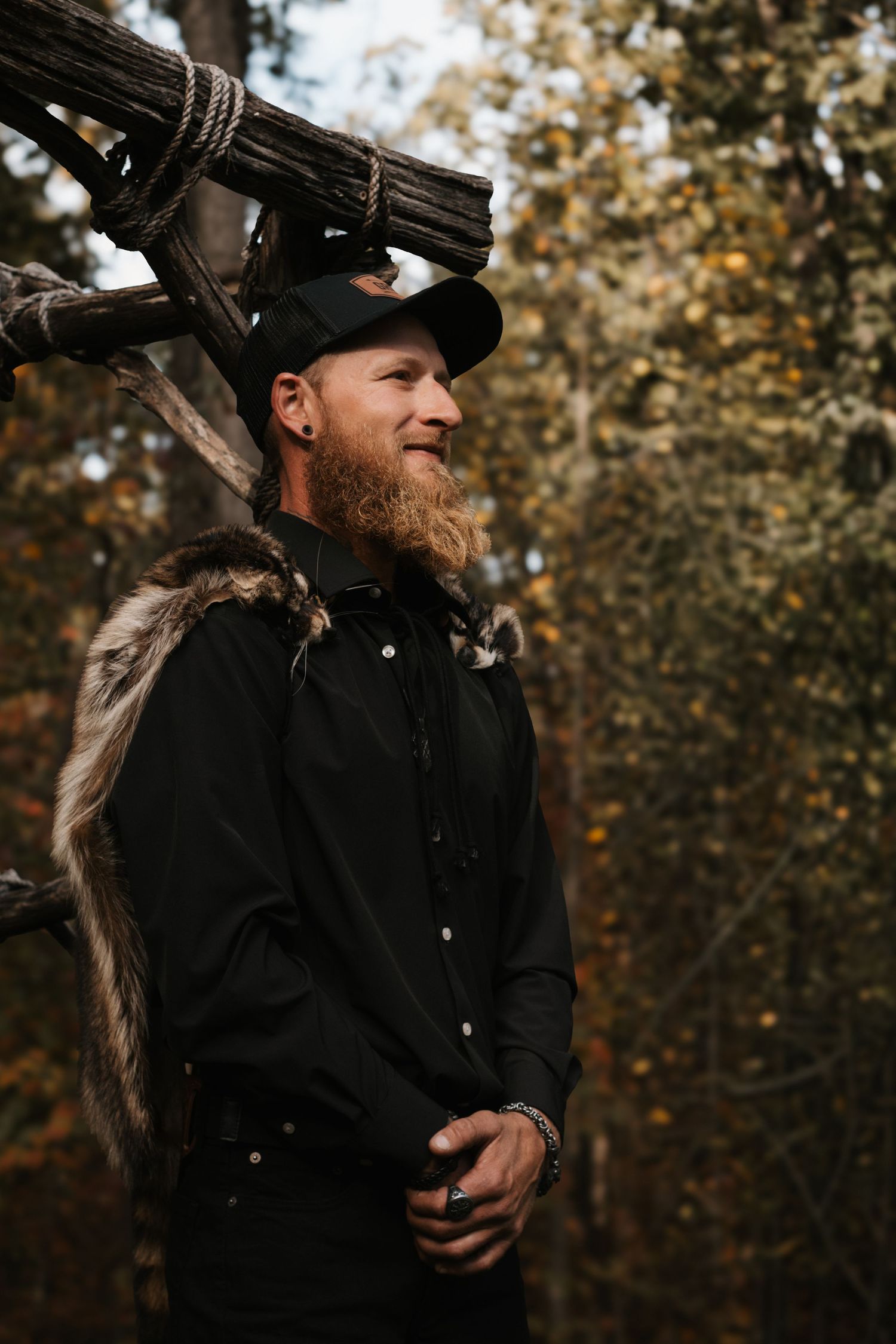 A person in a black hat and shirt smiles while standing against an autumn forest backdrop.