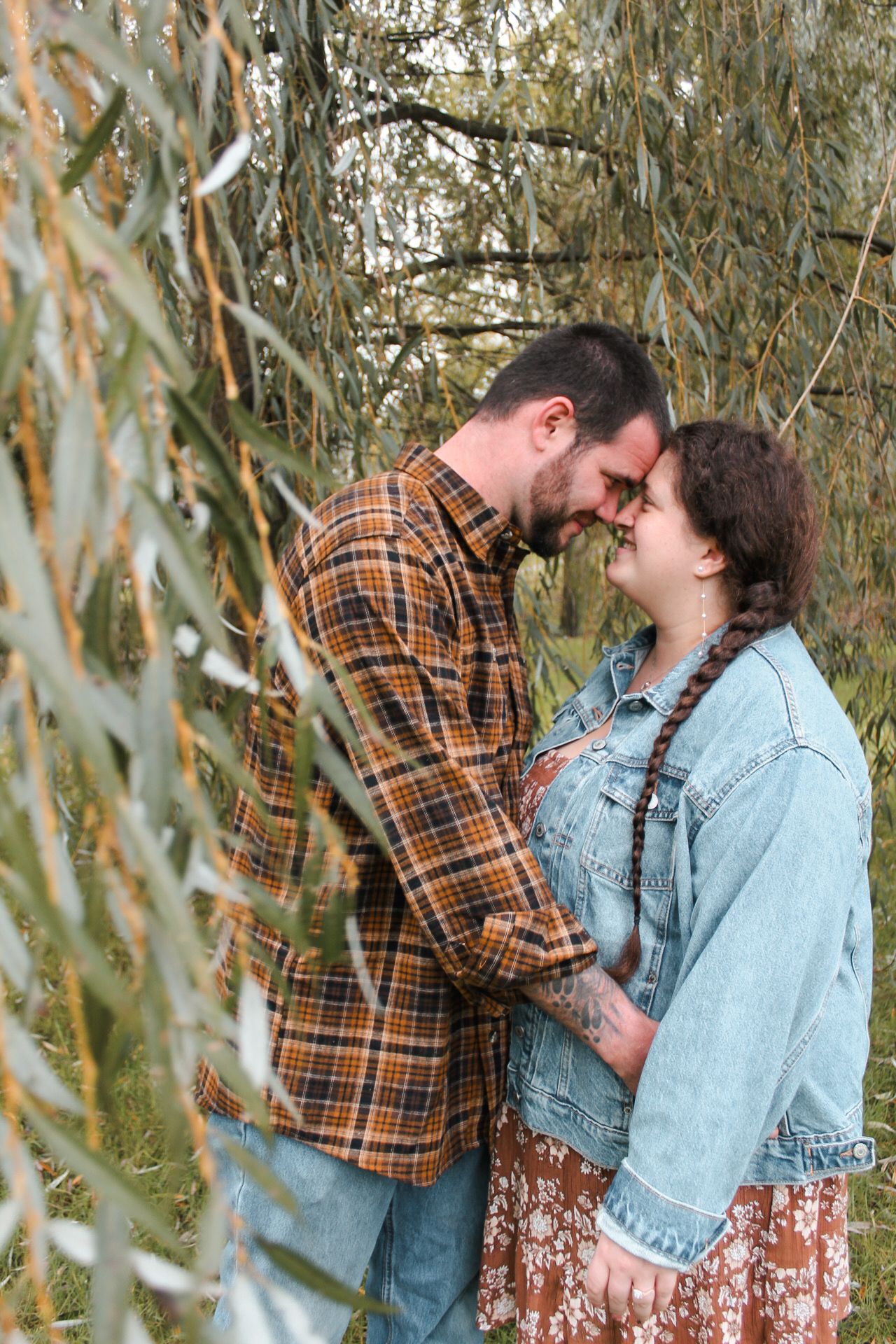 Romantic couple embracing under willow tree branches in casual fall attire.