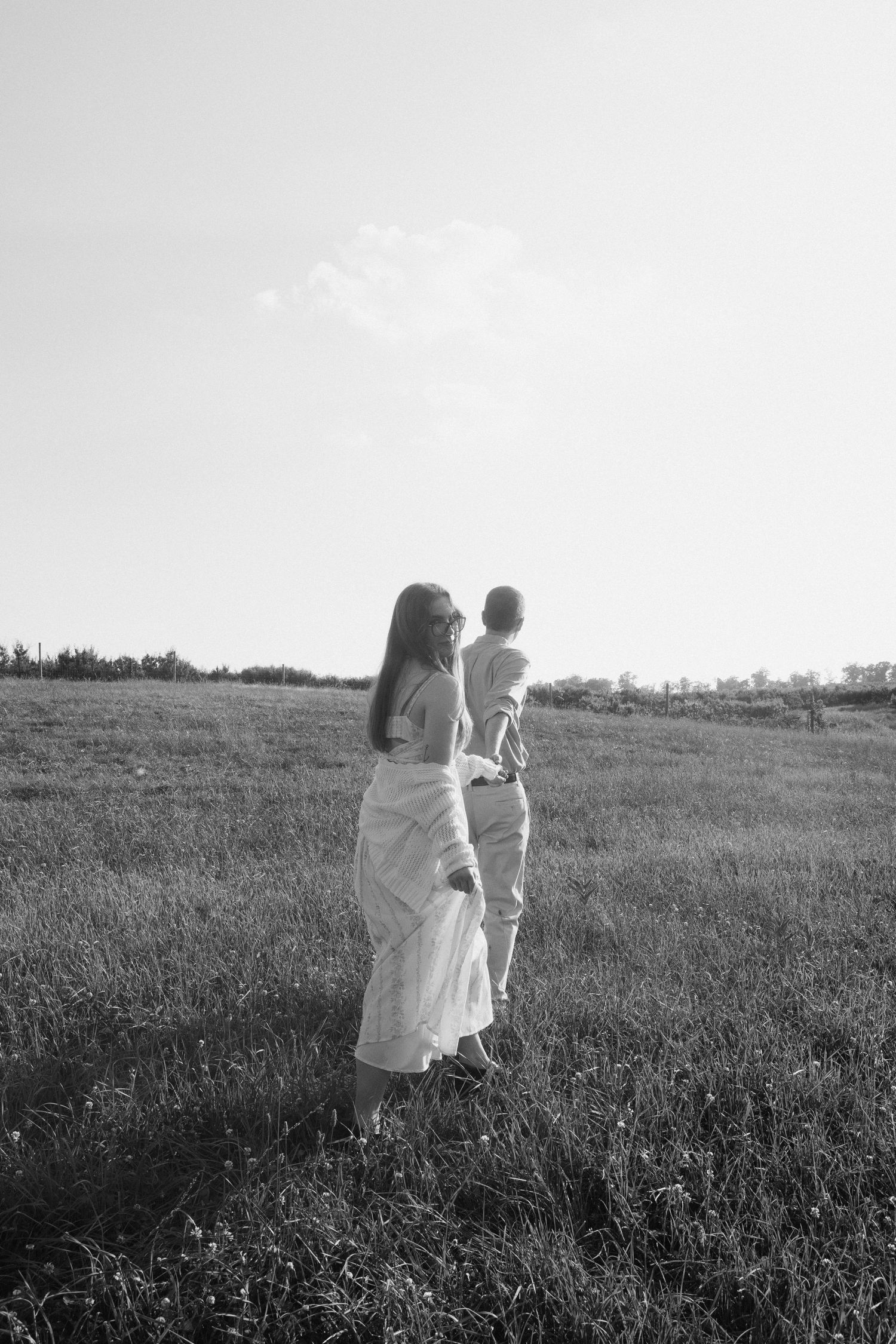 Black and white photo of couple standing in open field, woman in white dress looking back at camera.