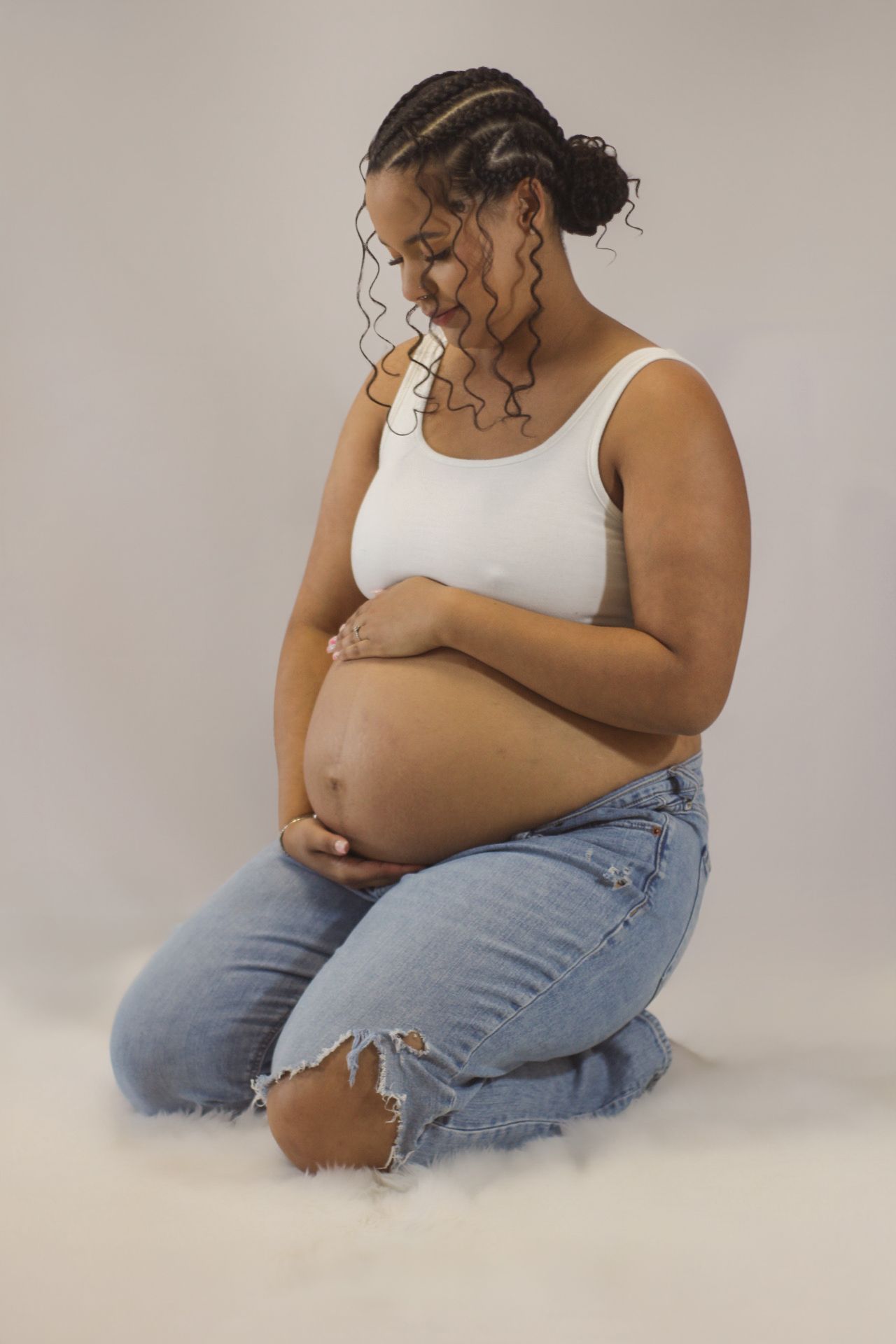 Tender maternity portrait of pregnant person in white tank top and jeans touching belly.