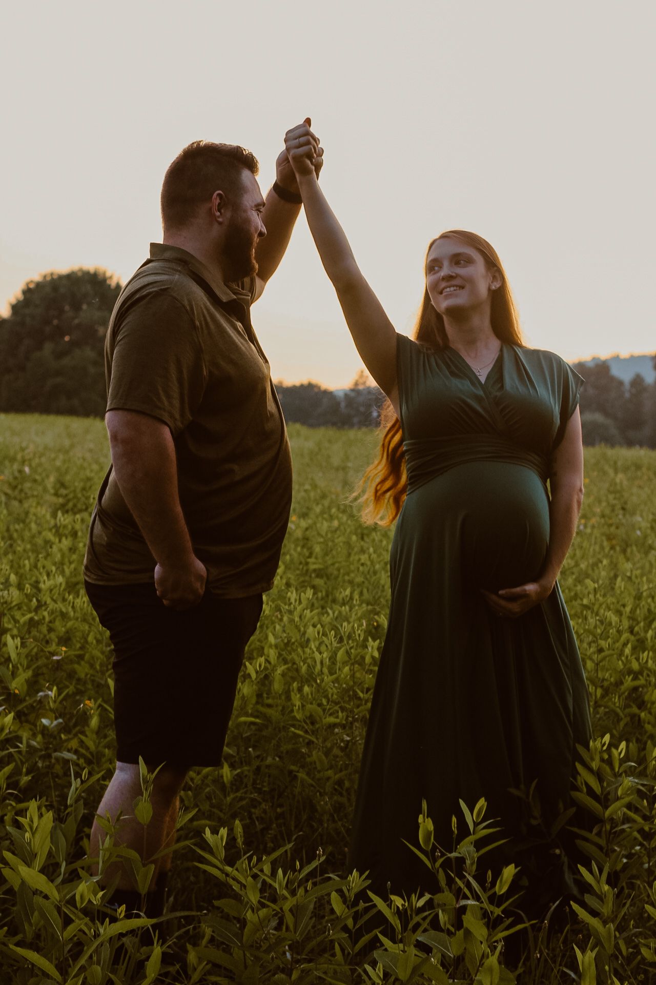 A couple dances in a field at sunset during a maternity photoshoot.