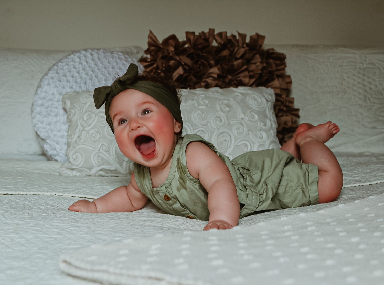 A baby in sage green clothing laughs joyfully while lying on white textured bedding.