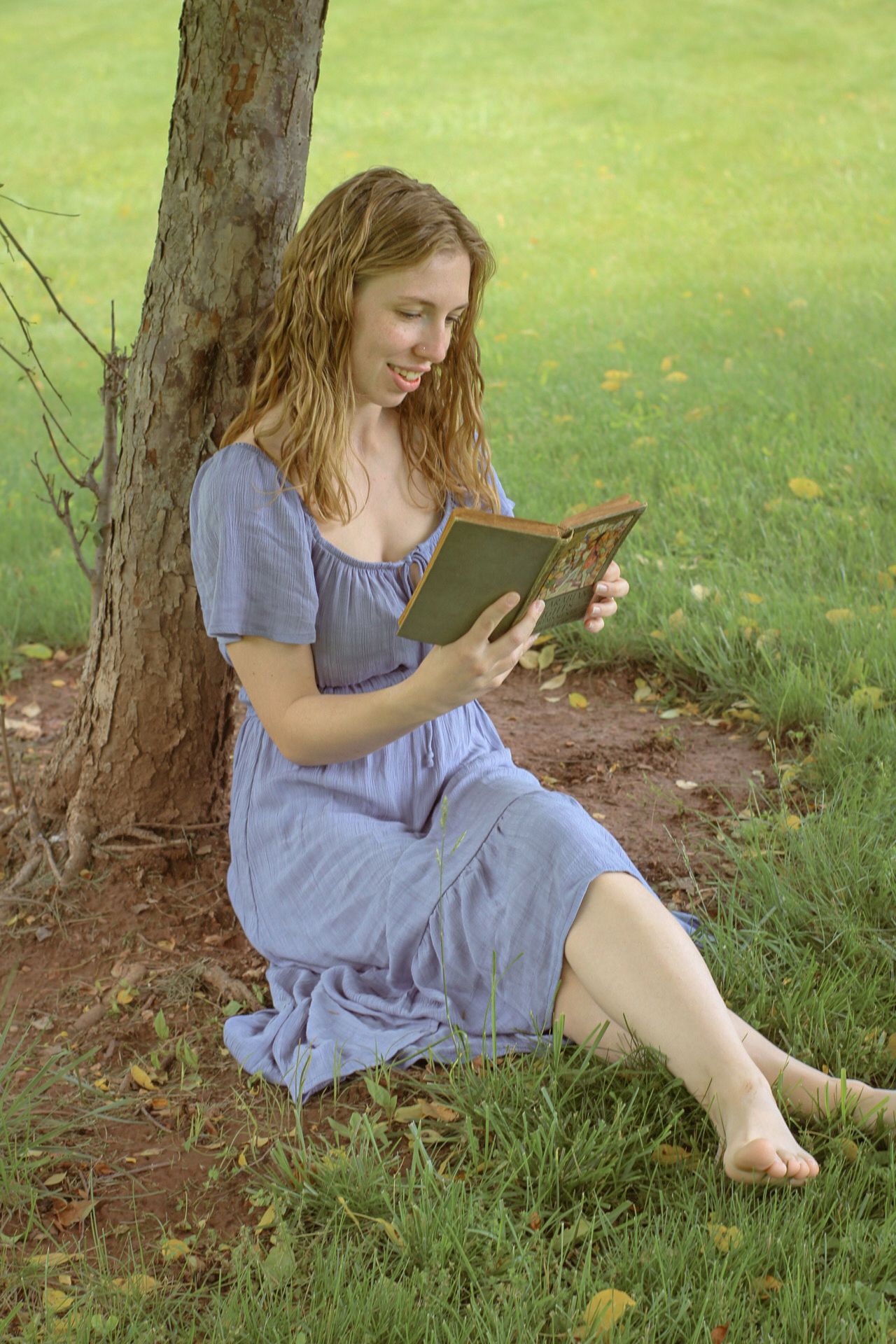 A person in a blue dress sits under a tree reading a book on a sunny summer day in the grass. Cottage core vibes, alice in wonderland vibes