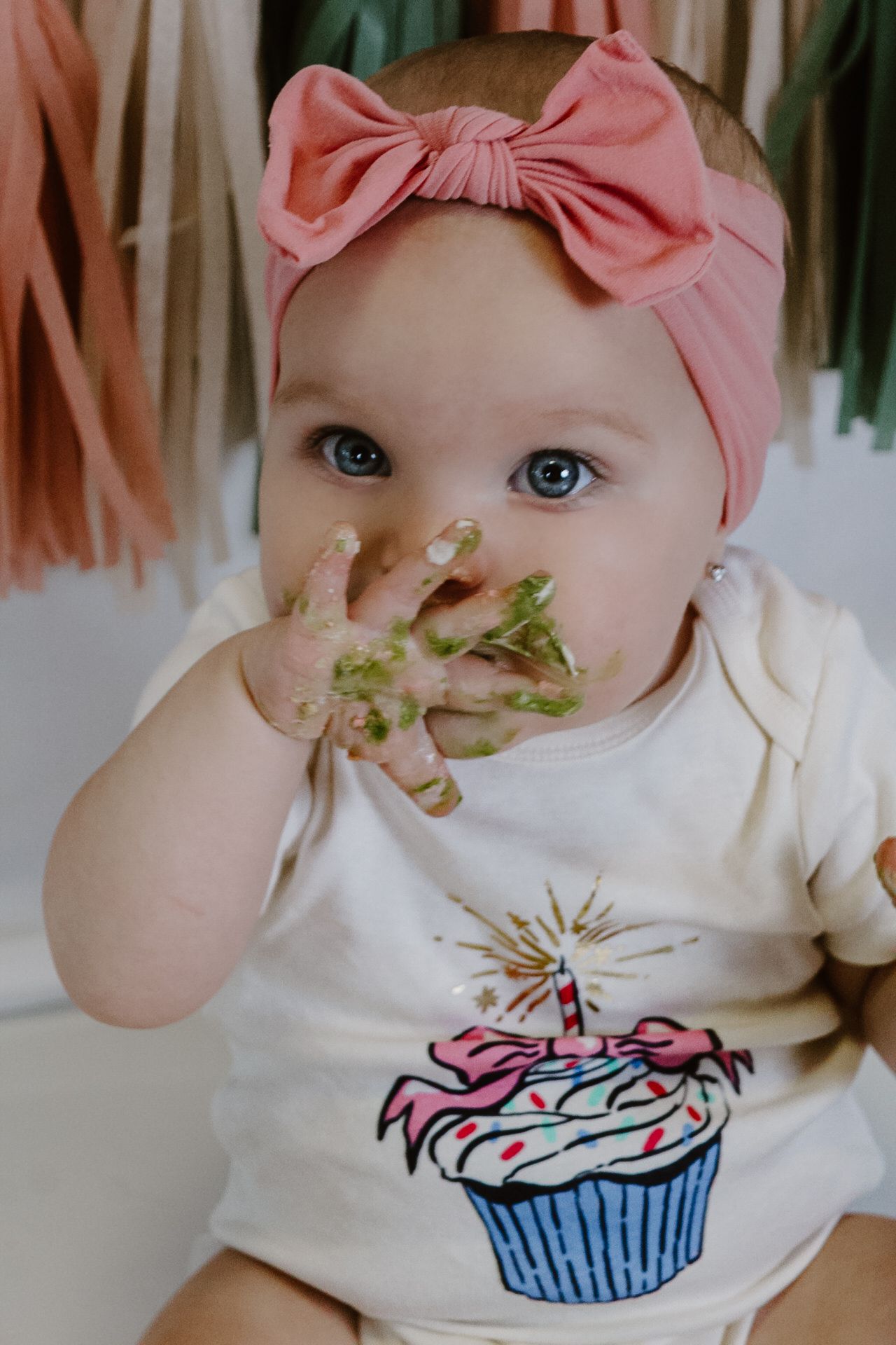 Cake smash photoshoot featuring cupcake onesie and pink bow against tassel backdrop.