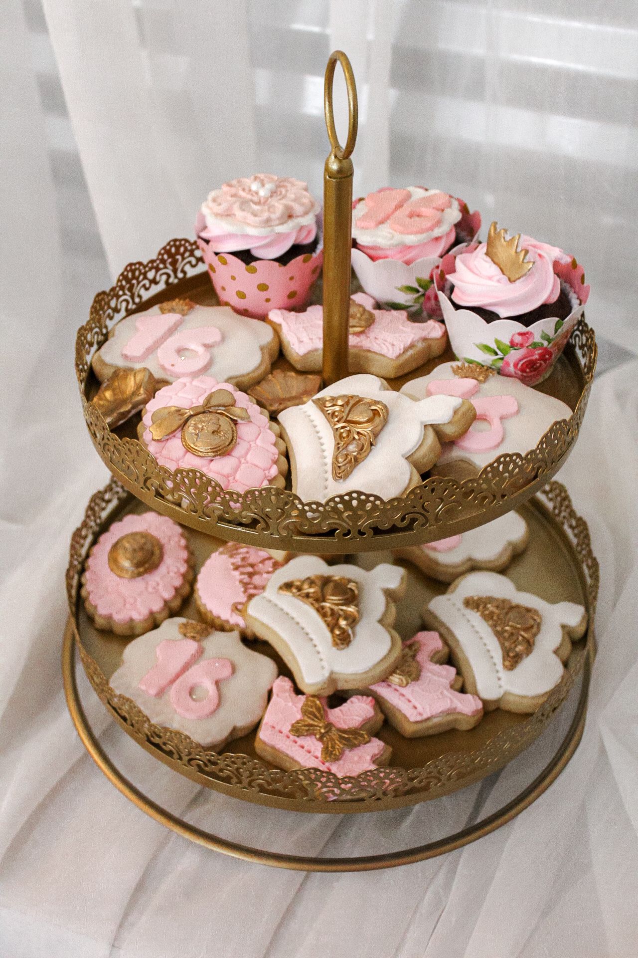 Two-tiered gold stand displaying pink and white decorated cookies and cupcakes.