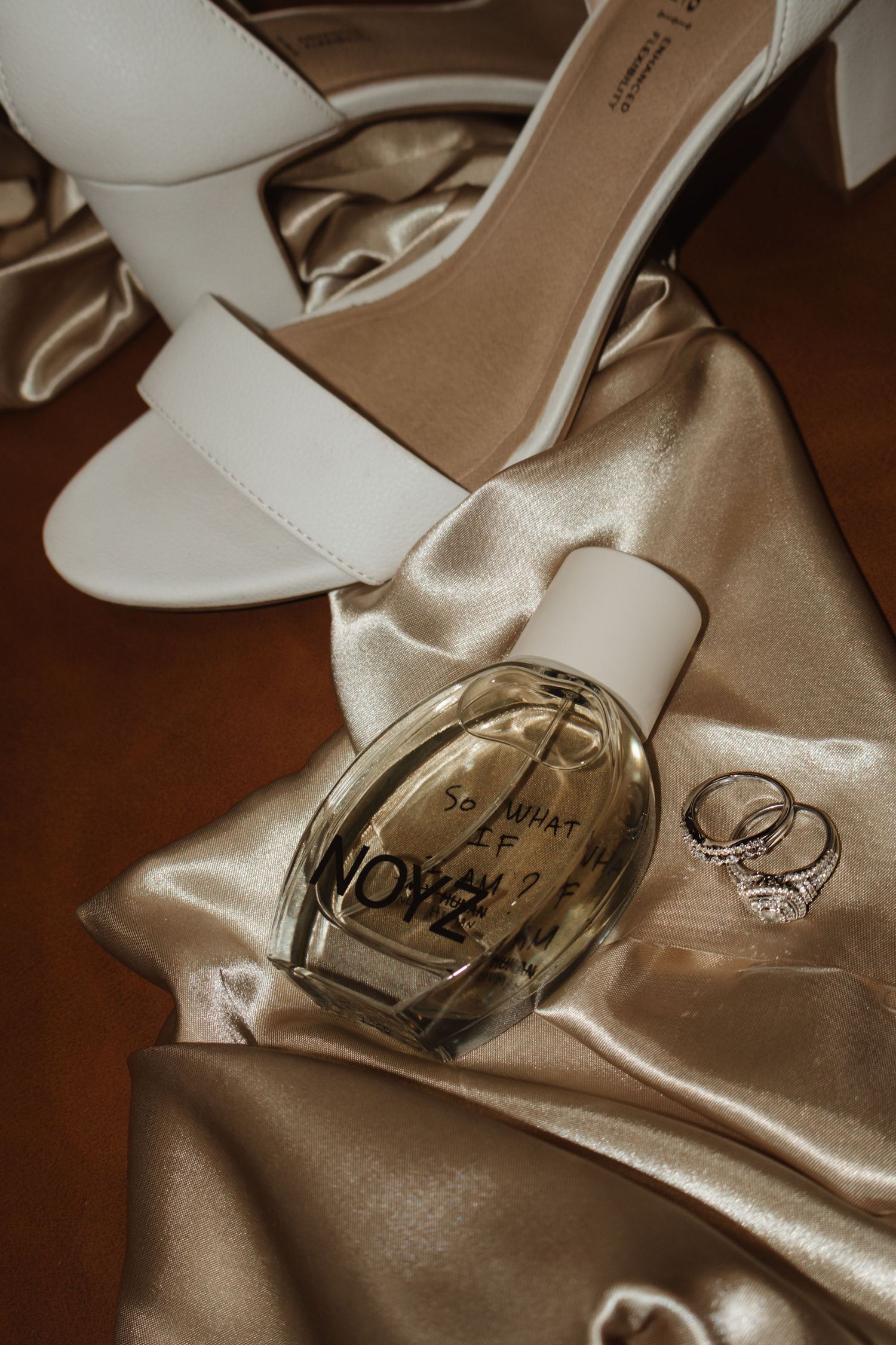 White bridal heels, perfume bottle, and diamond rings arranged on golden satin fabric for a wedding flat lay.