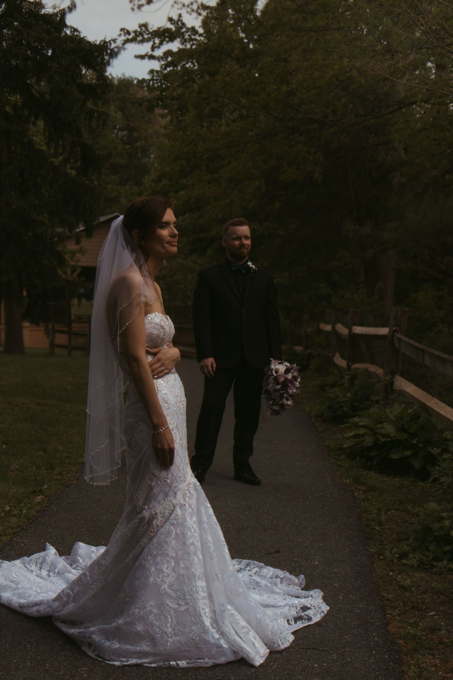 Bride in lace mermaid gown and veil stands on garden path with groom in black suit at dusk.