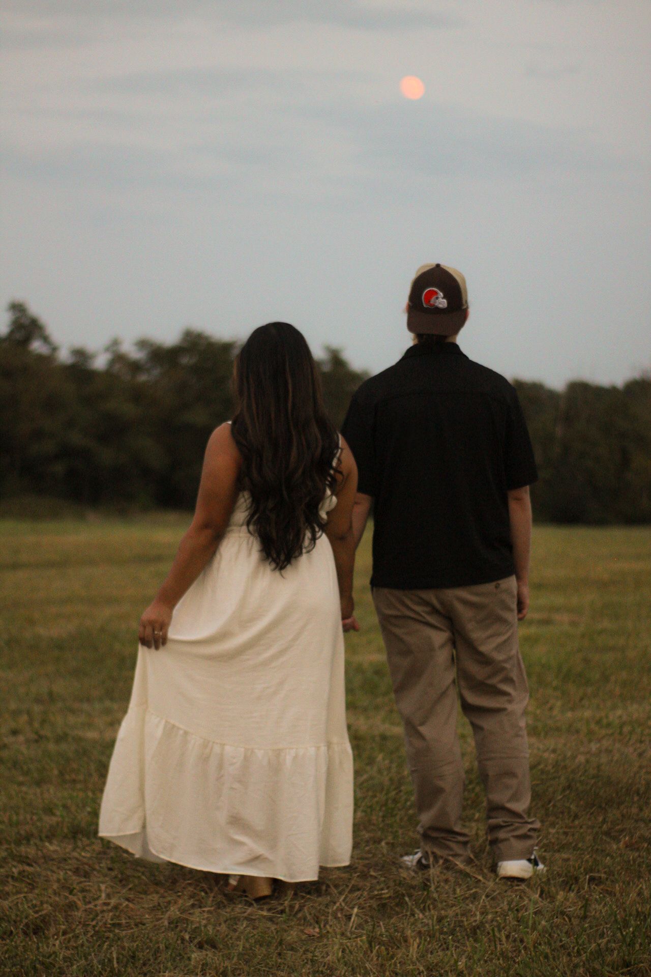 A couple stands together in a field at sunset, with the woman in a white dress and man in dark clothing.