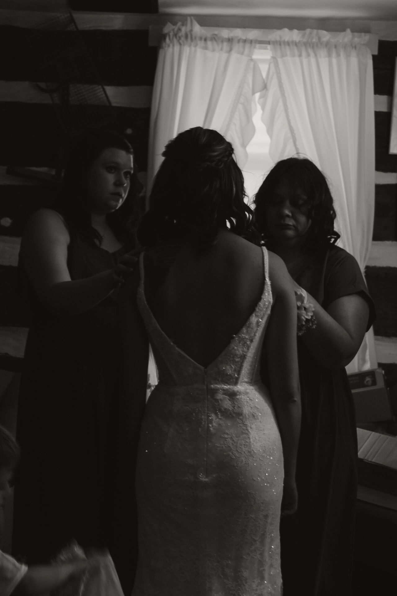 A bride in a white dress gets ready in a dimly lit room with curtains.