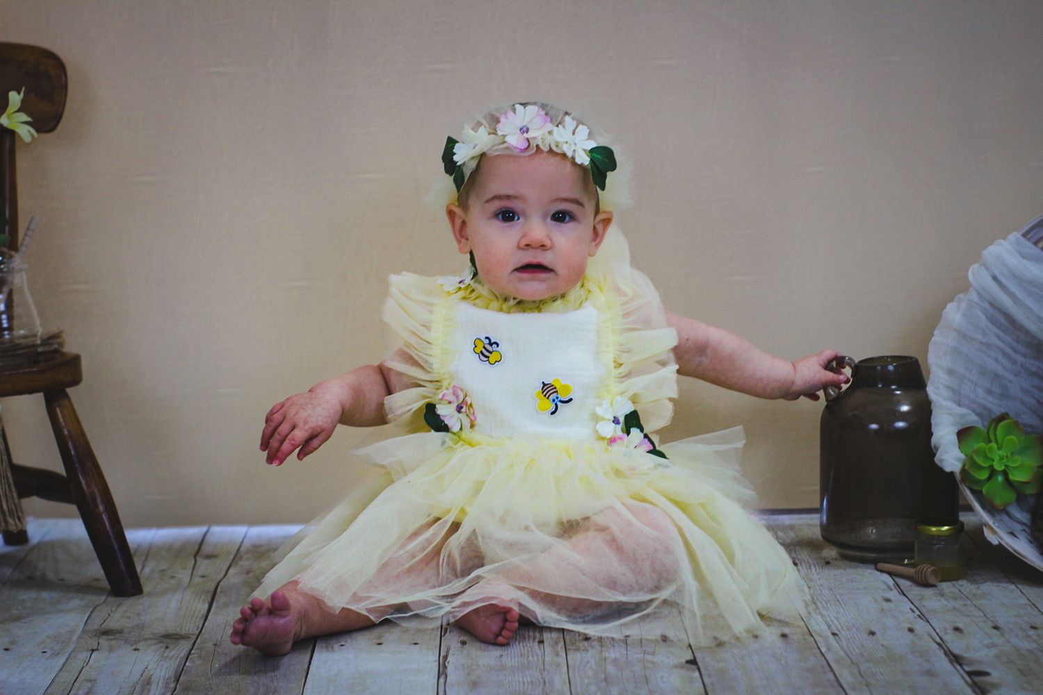 Baby wearing yellow sunflower dress and floral crown sitting on wooden floor.