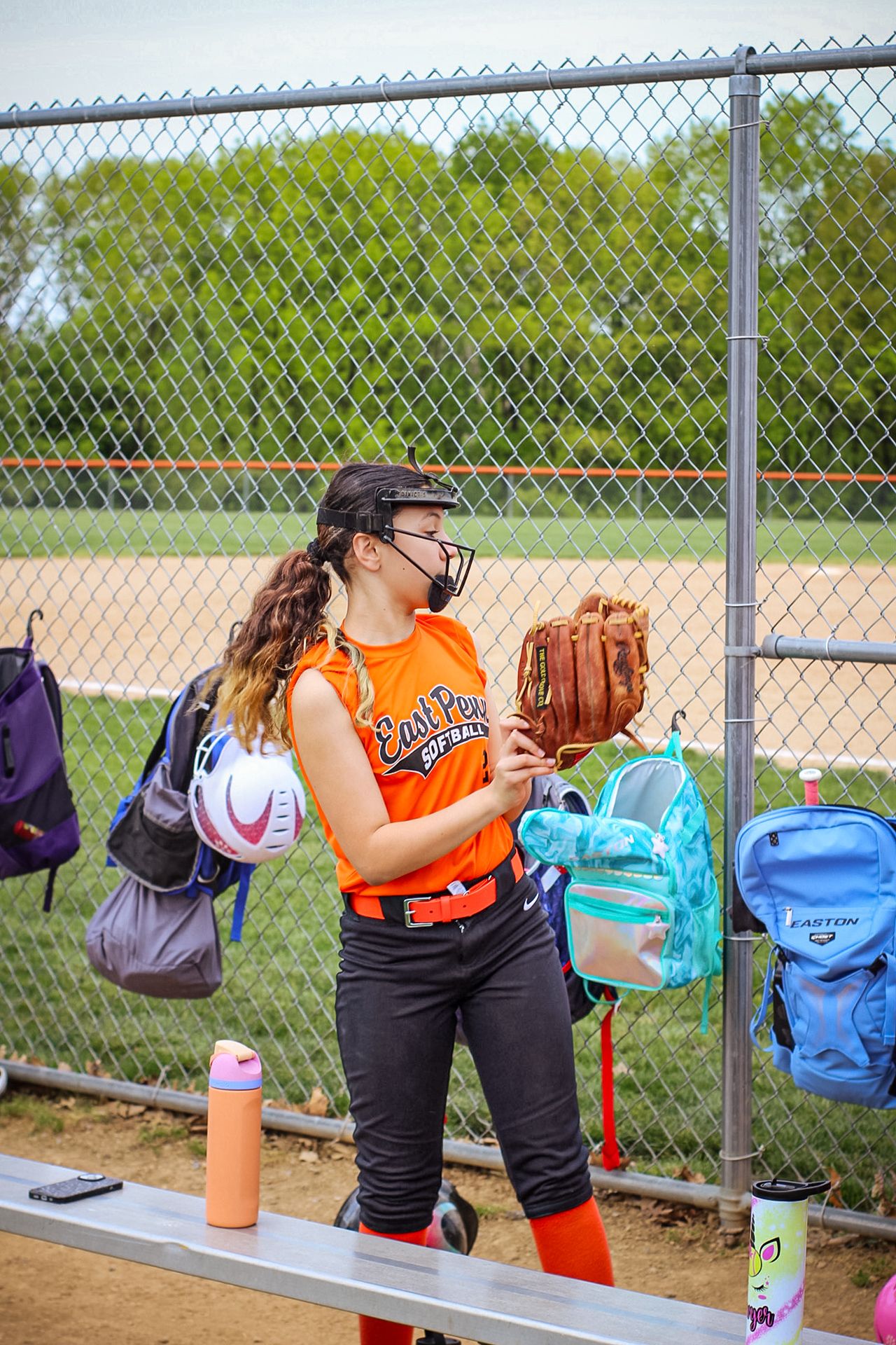 Softball player in orange jersey by dugout fence with colorful backpacks hanging.