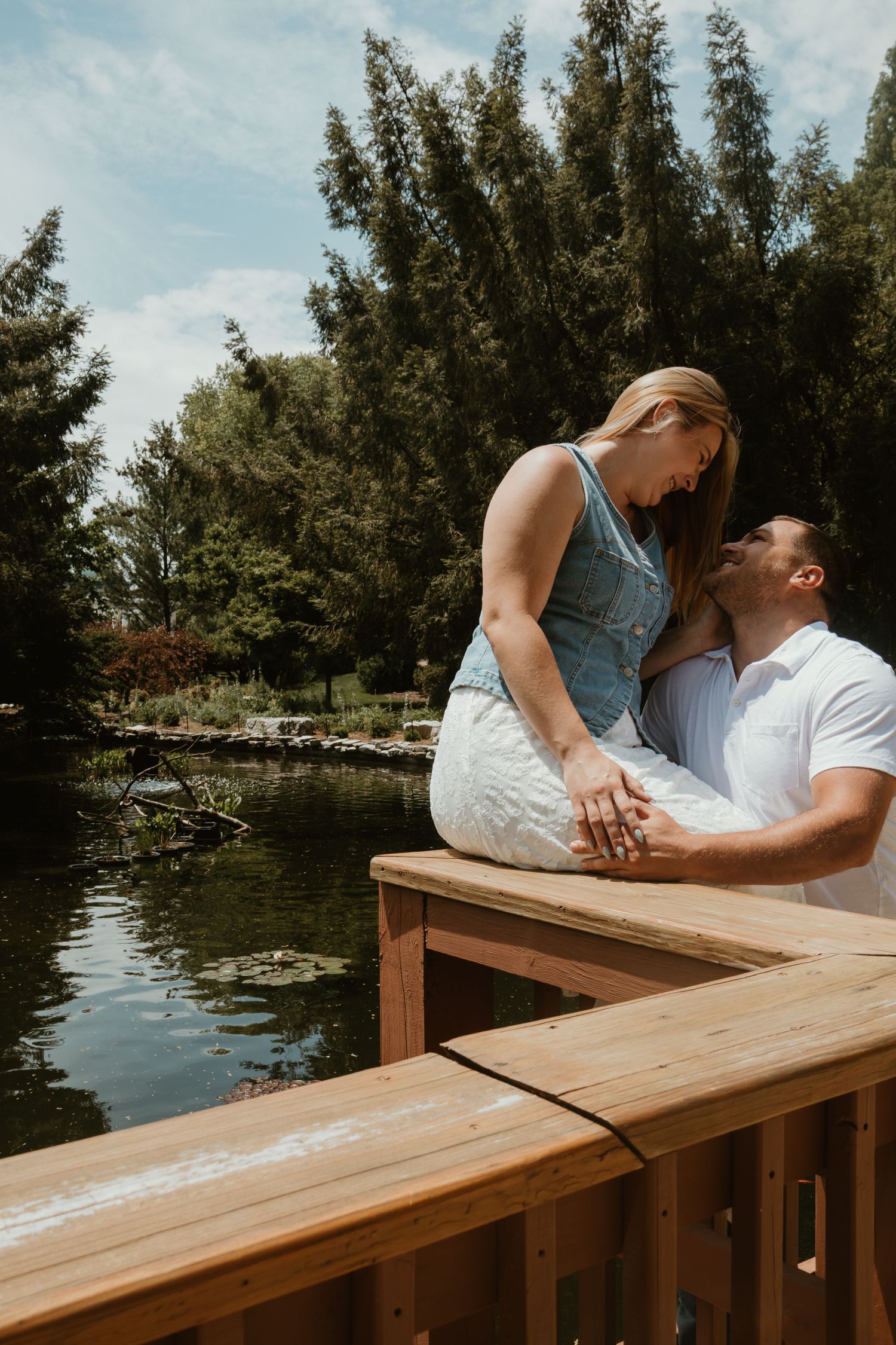 Couple sharing a romantic moment on a wooden dock beside a serene pond in a lush green park on a sunny day.