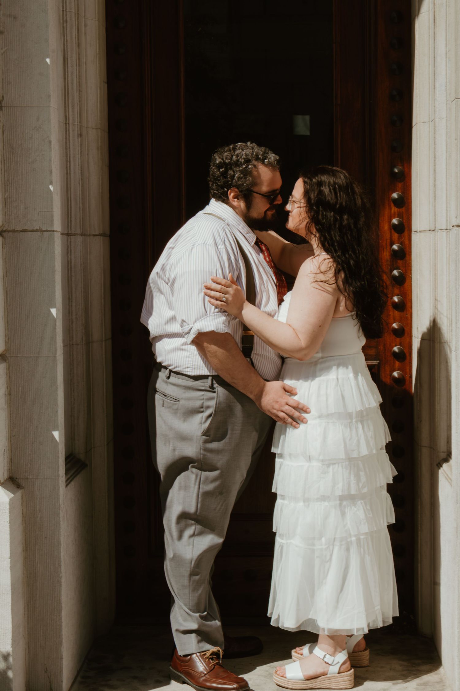 Bride and groom sharing a kiss in a grand doorway, bride in white ruffled dress, groom in gray trousers.