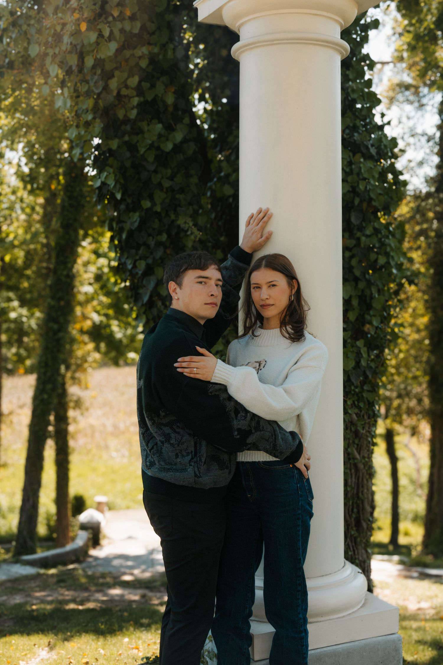 Young couple posing by a white column in a lush green park, embracing in casual autumn outfits.