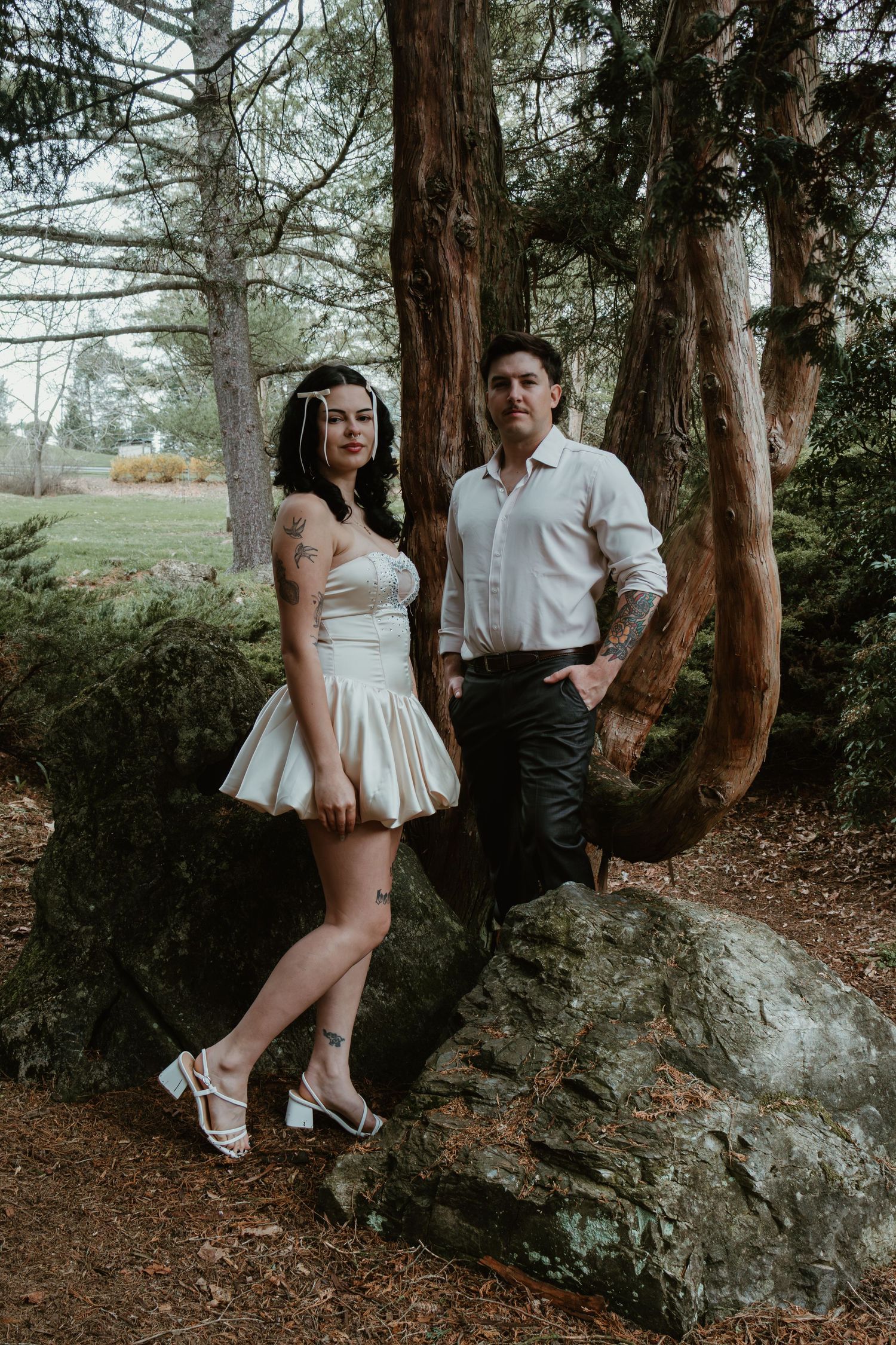 A couple poses among mossy rocks and tall trees, woman in white mini dress and man in white shirt and dark pants.