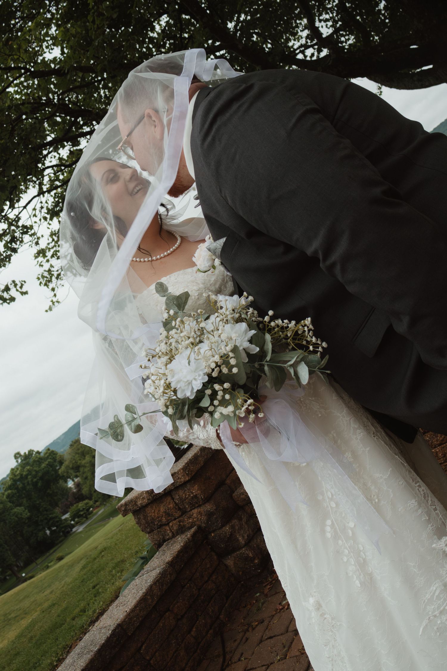 Bride and groom share a romantic kiss under a veil outdoors, bride holding a white floral bouquet.