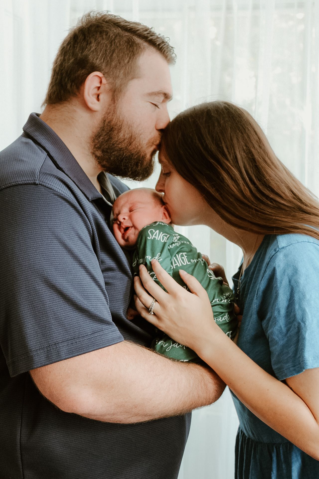A couple shares an intimate moment with their newborn baby wrapped in a green swaddle blanket.