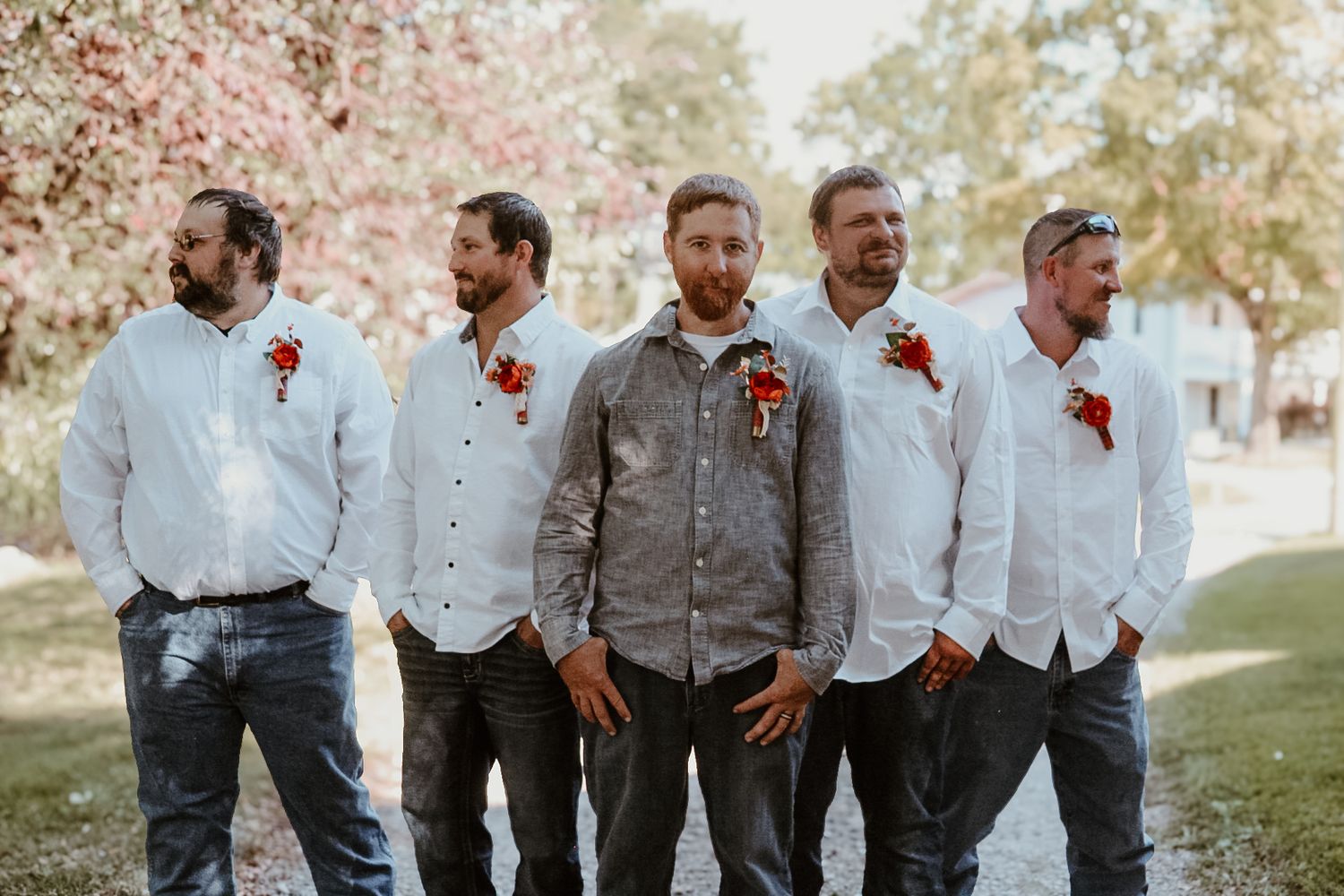 Groomsmen in light gray suits and white shirts pose together with red boutonnieres outdoors.