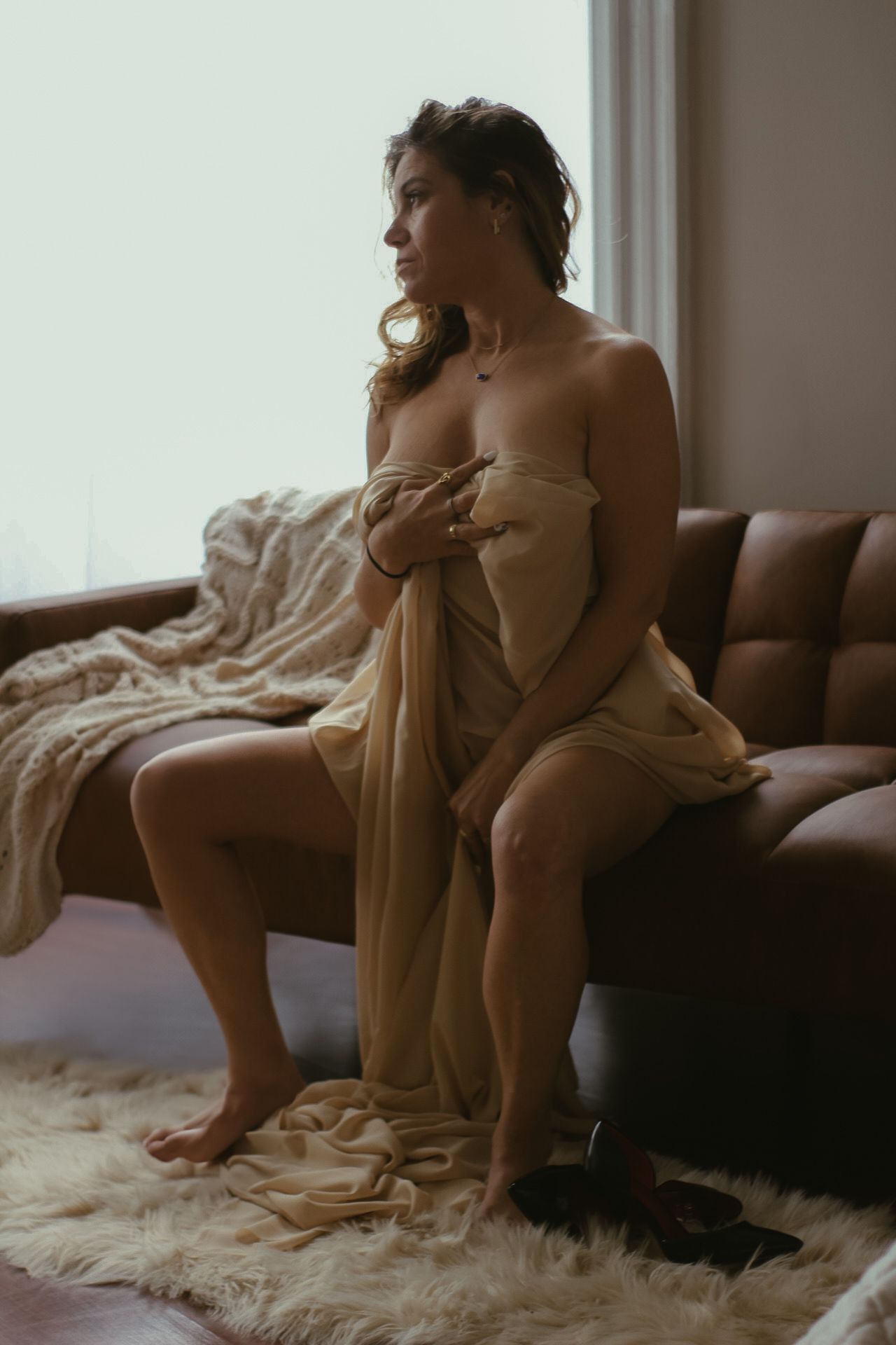 Artistic boudoir photograph in moody natural lighting of someone draped in beige fabric sitting on brown leather couch.