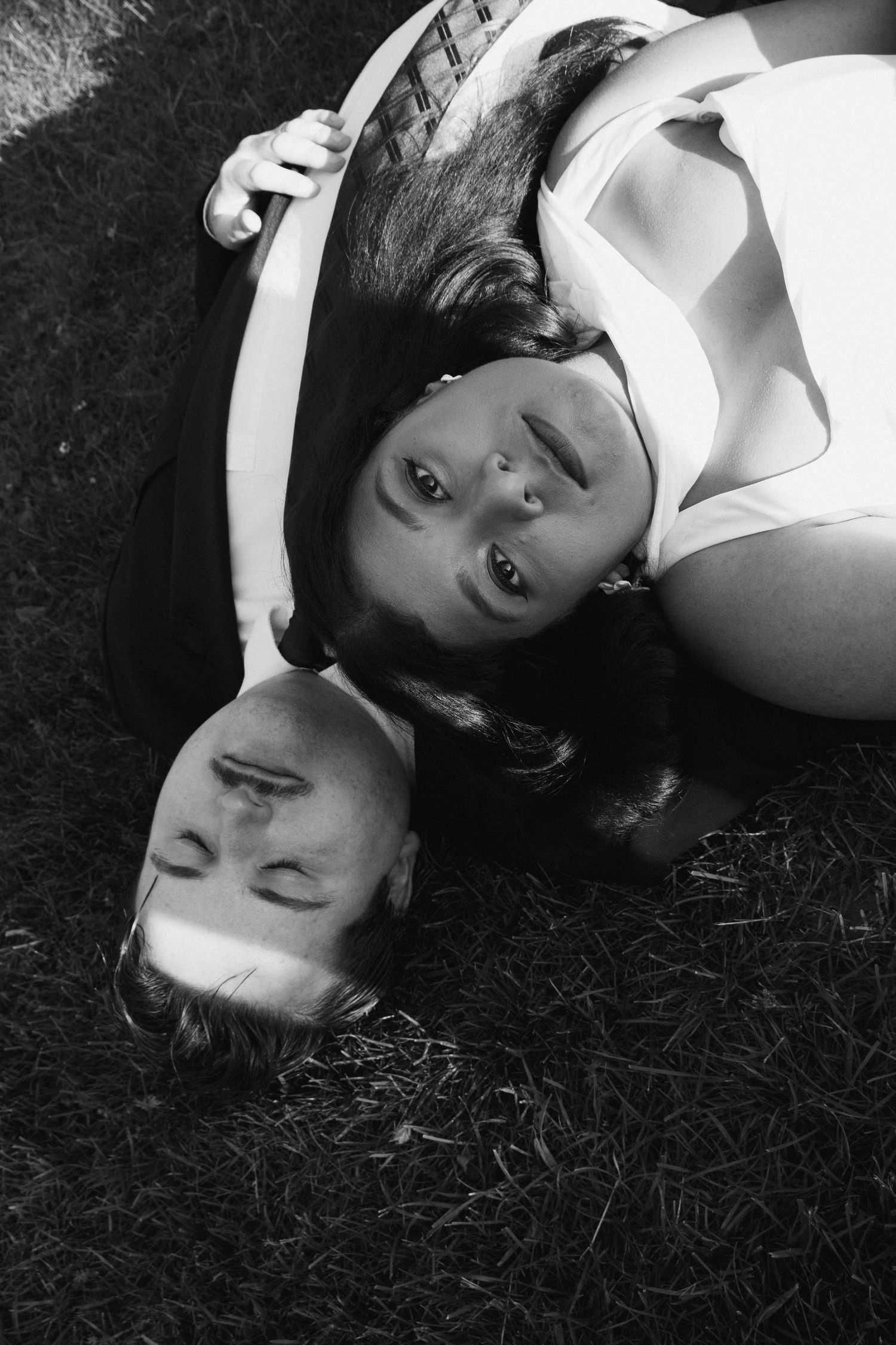 Black and white photo of a couple lying on grass, heads together, smiling up at the camera.