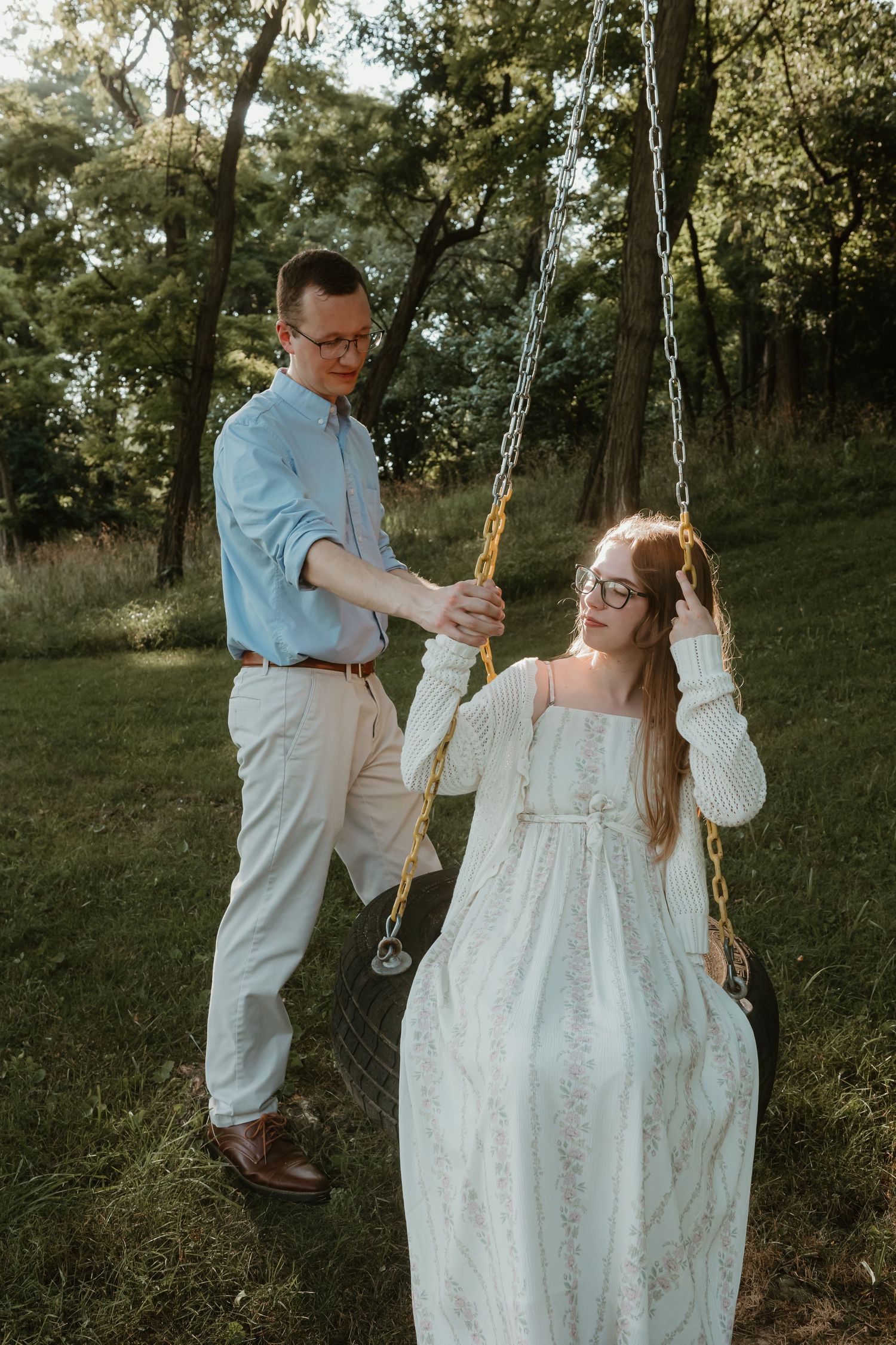 A man pushes a woman in a white dress on a tire swing outdoors in a lush green garden setting.