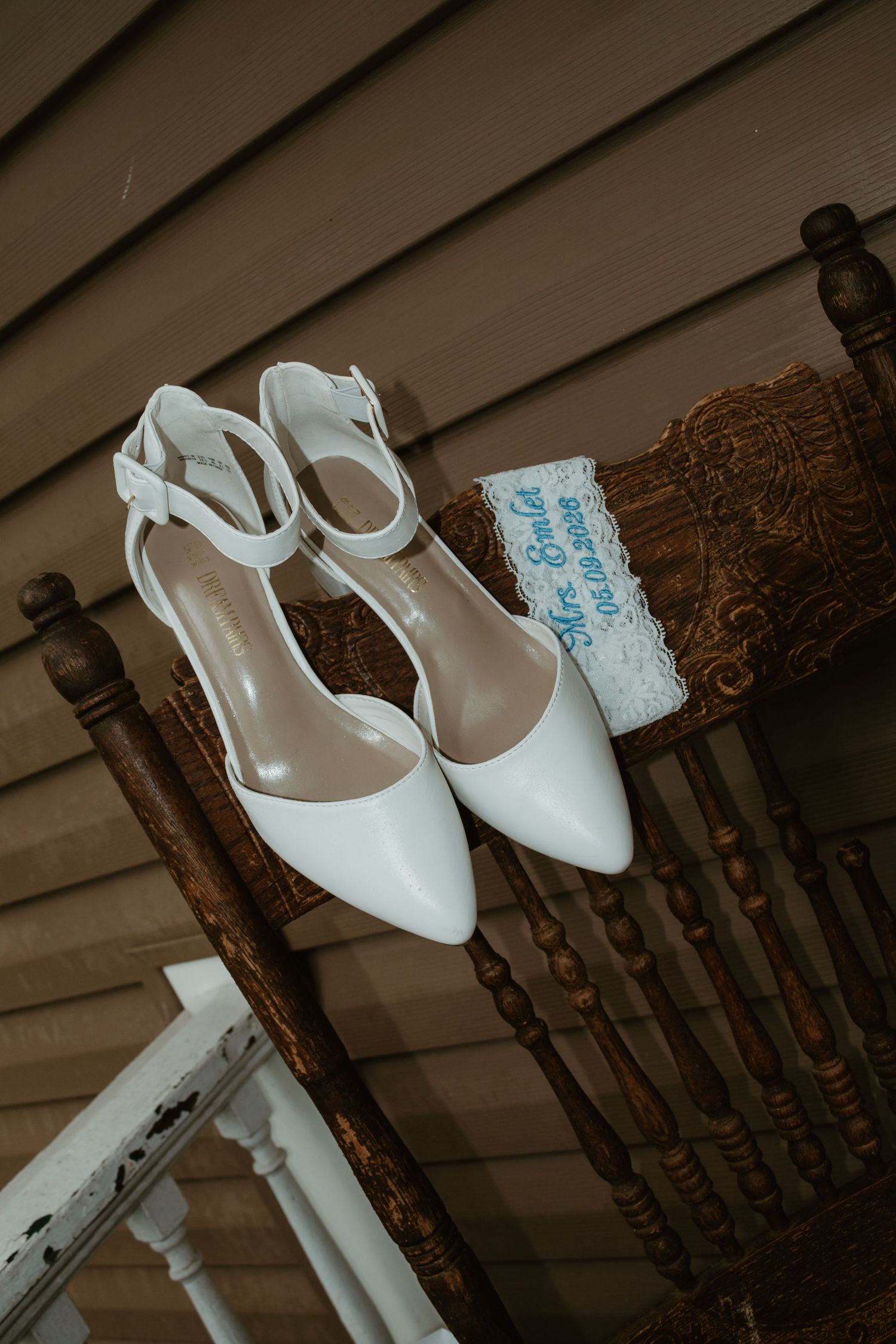 White bridal heels with ankle straps displayed on a wooden chair beside a lace wedding garter with blue embroidery.