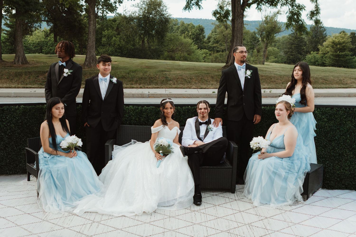 Wedding party poses outdoors, bride and groom seated center, flanked by bridesmaids in blue gowns and groomsmen in black tuxedos.