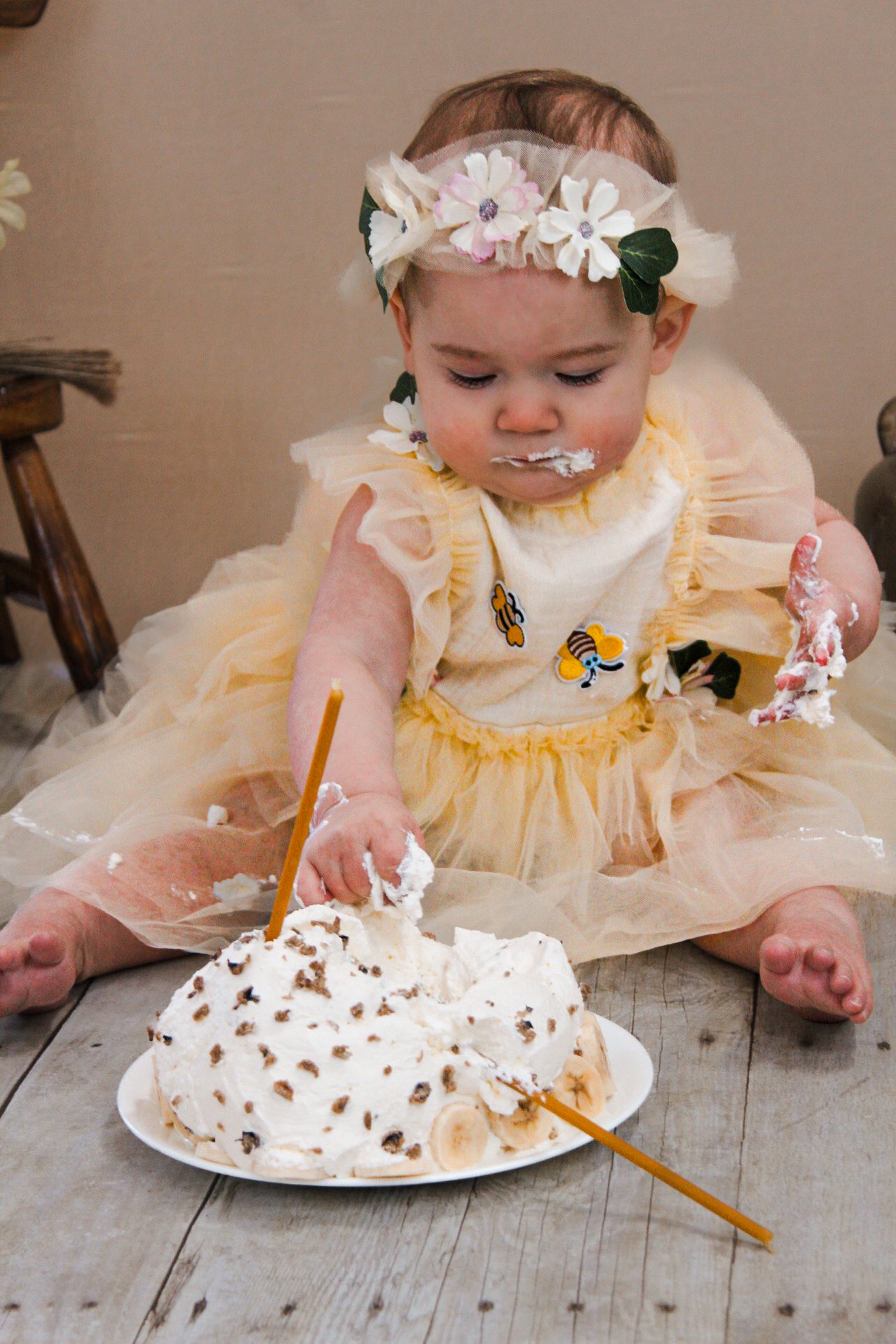 A baby in a yellow dress smashes into a white decorated cake on a wooden table.