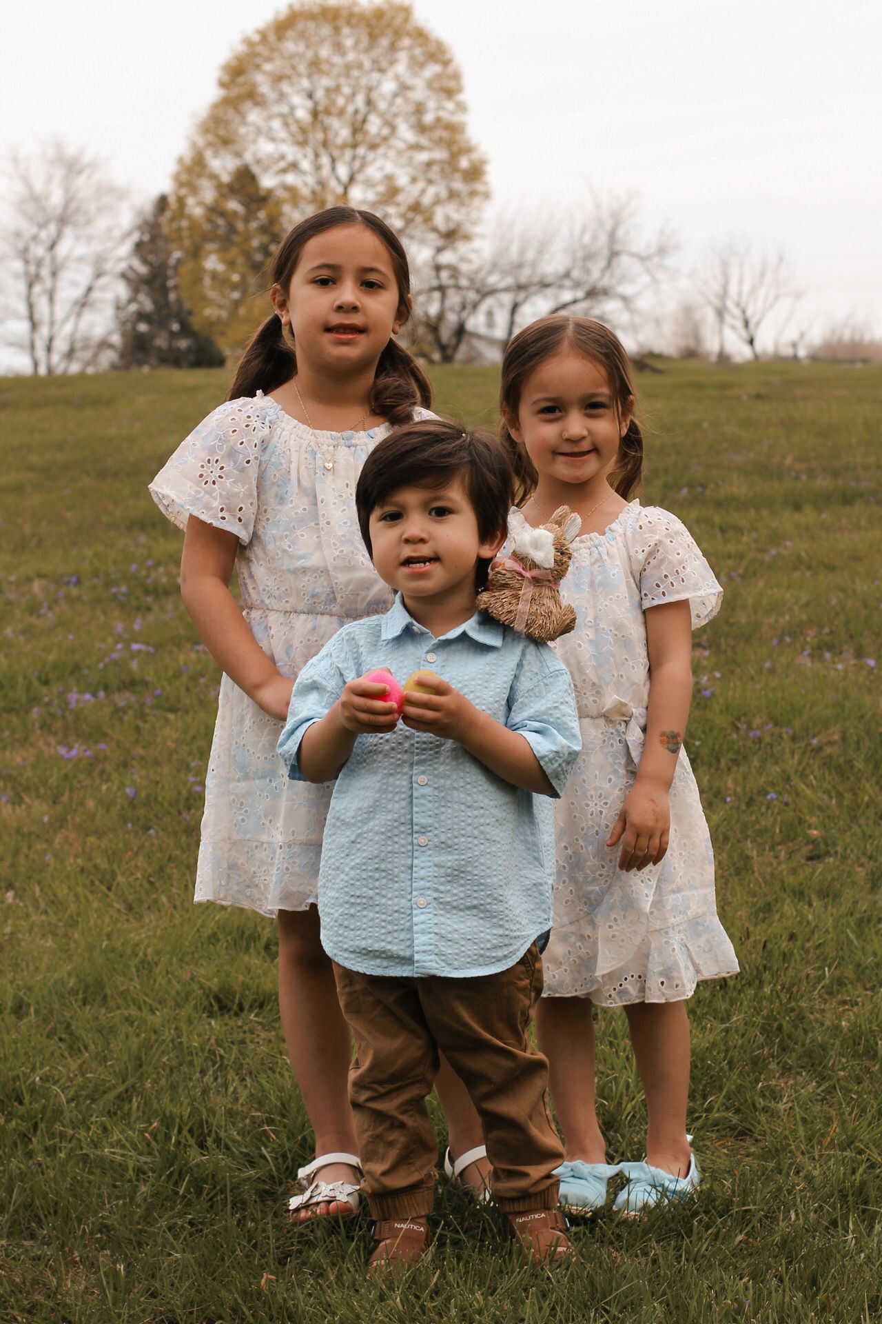 Children in white dresses and casual wear stand together in a grassy field during golden hour.