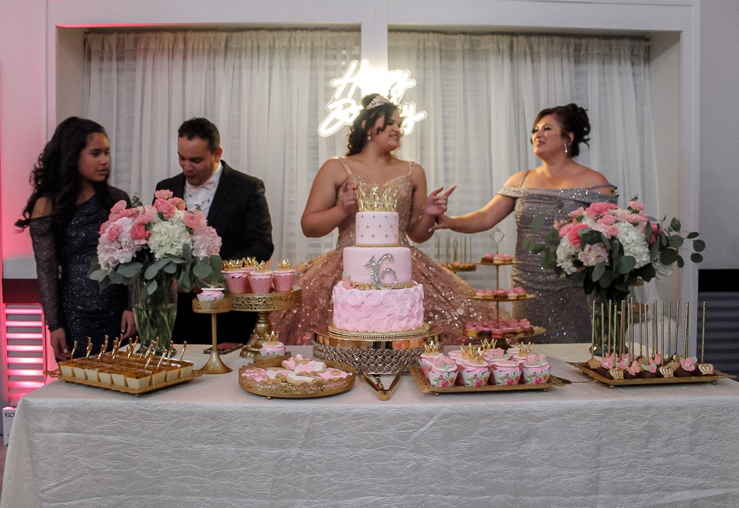 Elegant pink and white party setup with dessert table featuring cakes, flowers and decorative elements.