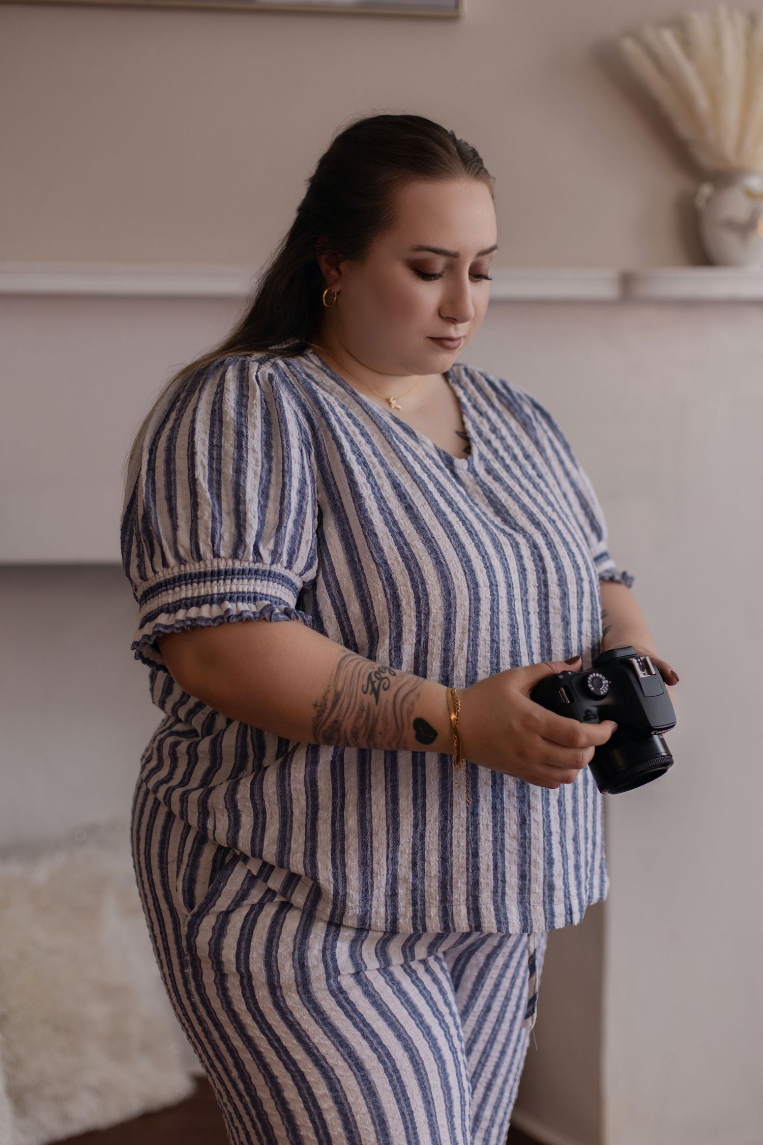 A photographer wearing a striped dress holds a professional camera while standing in a minimalist interior setting.