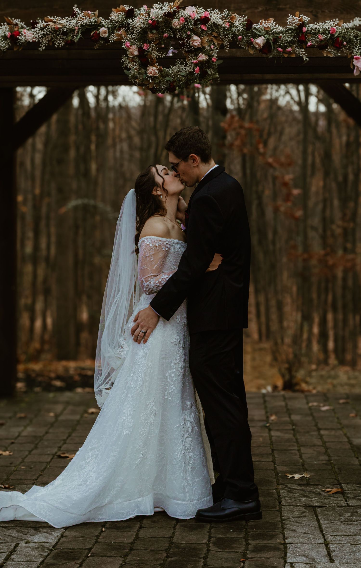 A romantic wedding couple embraces on a stone patio surrounded by autumn trees and hanging floral decorations.