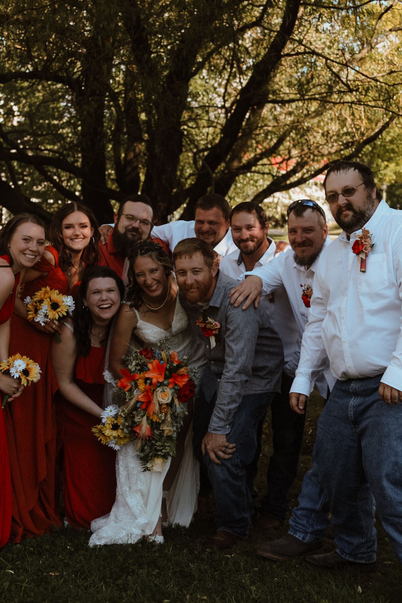Wedding party poses together under trees wearing burgundy dresses and casual attire for fun group photos.