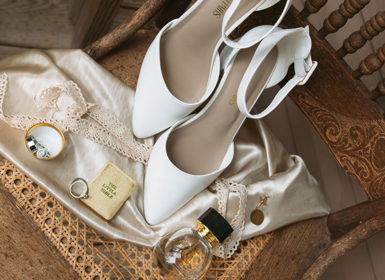 White bridal heels with ankle strap displayed on satin fabric alongside perfume, jewelry, and lace accessories.