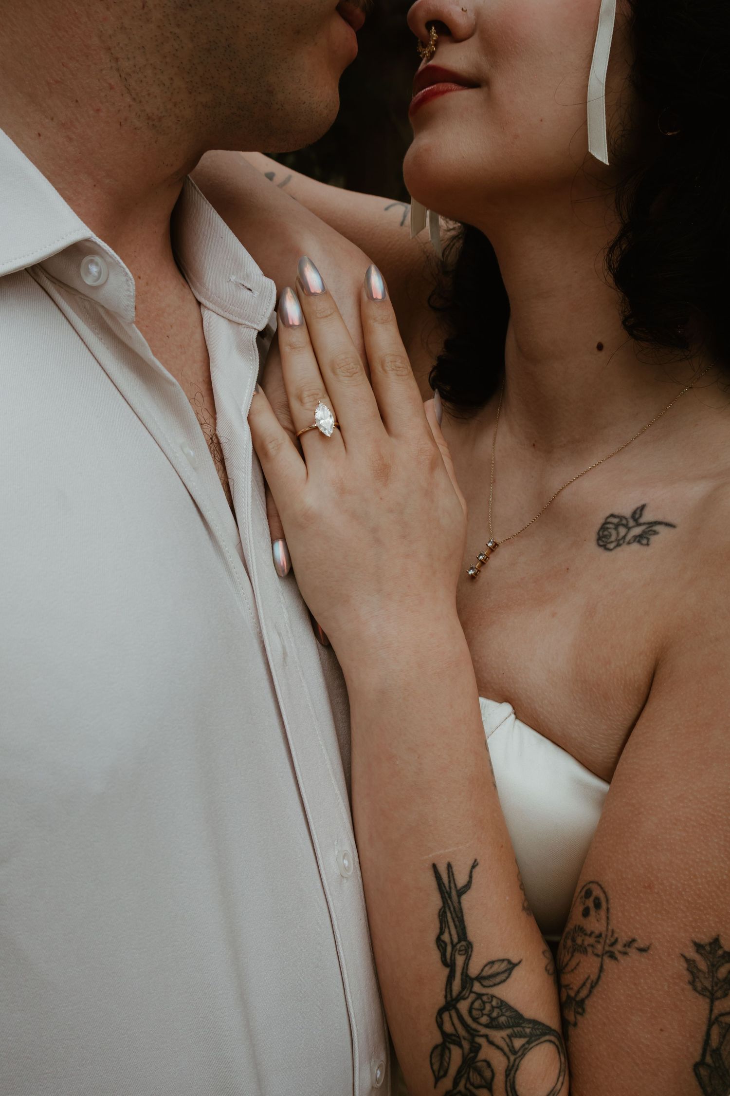 Tattooed bride with gold engagement ring rests hand on groom's chest in intimate wedding portrait.