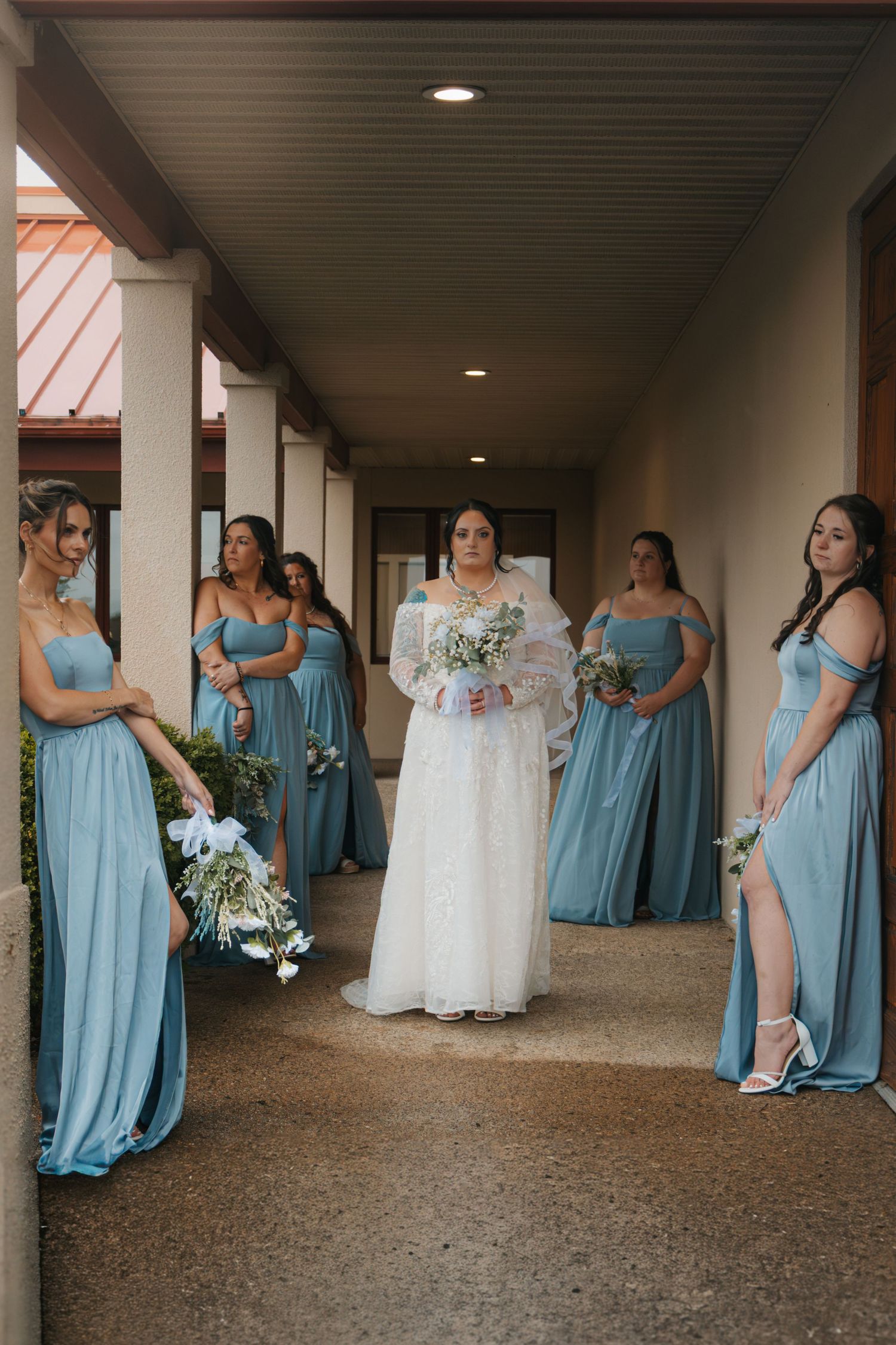 Bride in white gown stands with bridesmaids in blue dresses holding bouquets in a covered walkway.