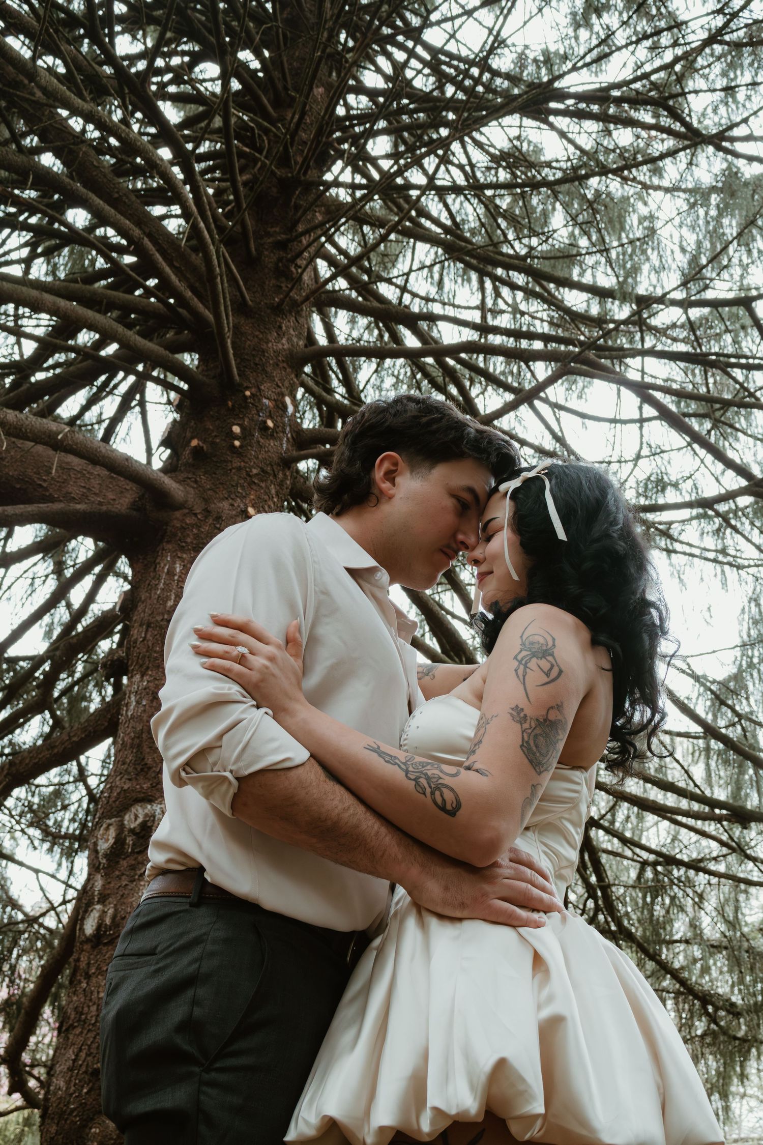 Tattooed couple in wedding attire sharing a romantic moment under a pine tree, bride in white dress with headband.
